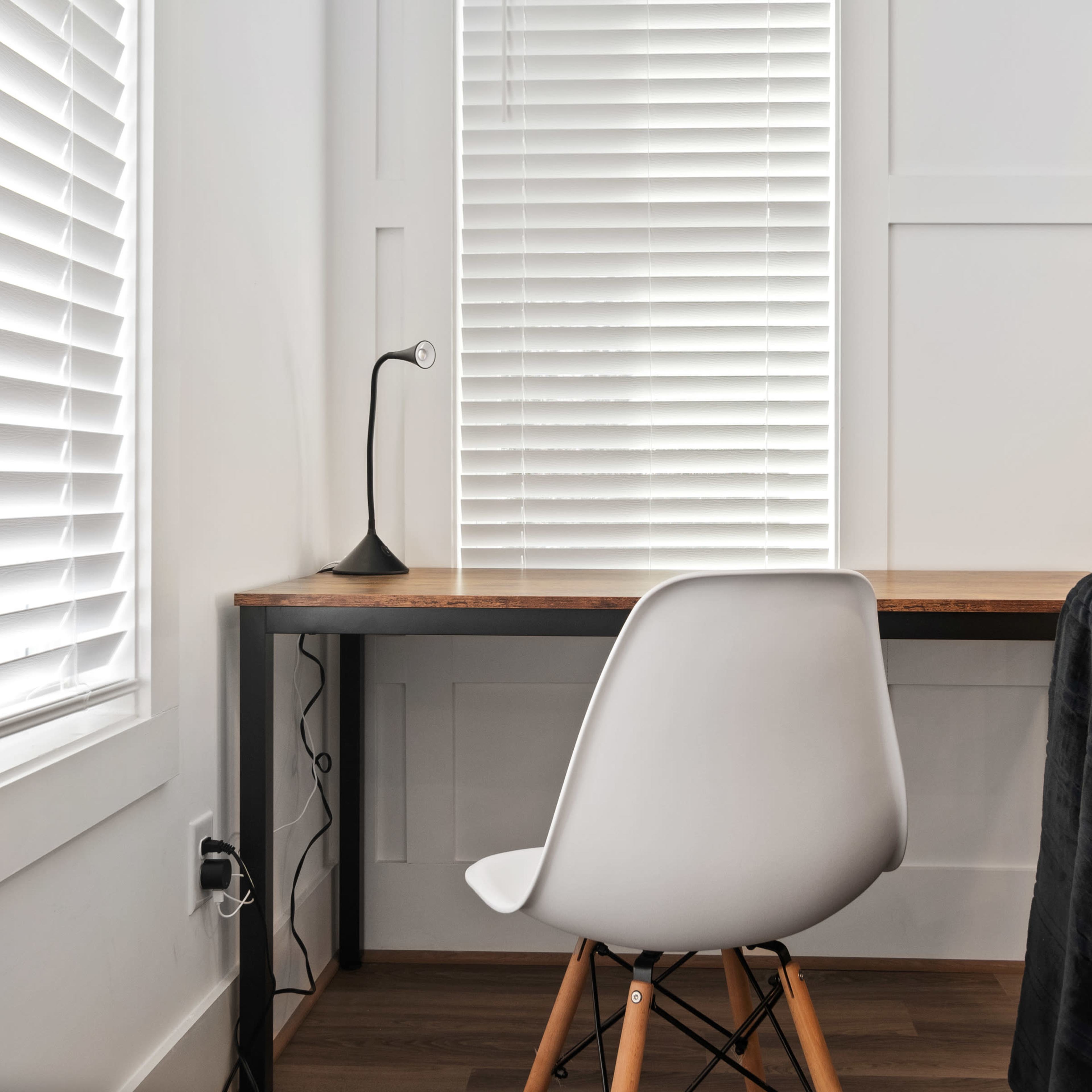 A minimalist workspace featuring a wooden desk, a white chair, and a desk lamp next to a window with blinds.