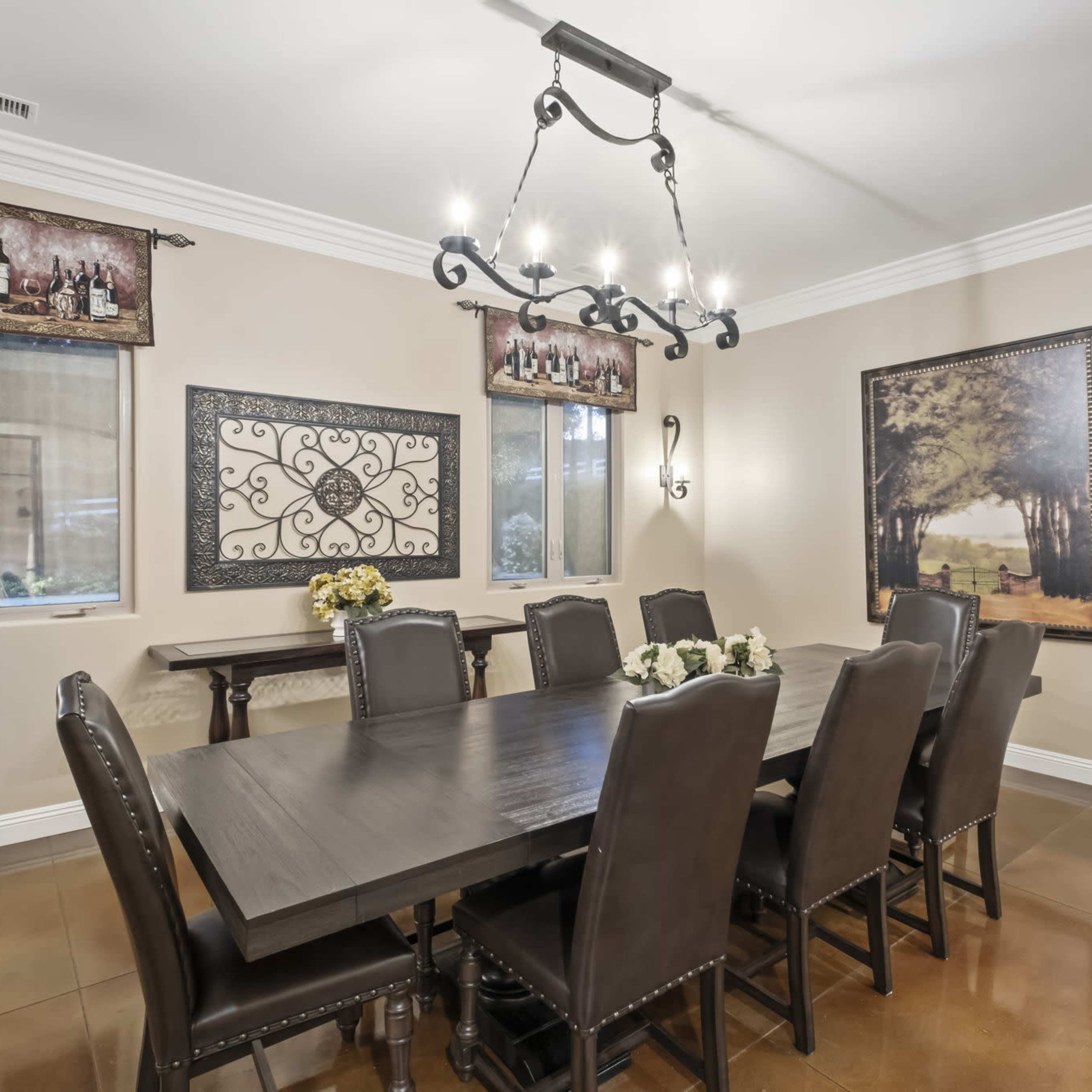 A dining room features a large wooden table surrounded by eight black chairs, with two windows and decorative artwork on the walls.