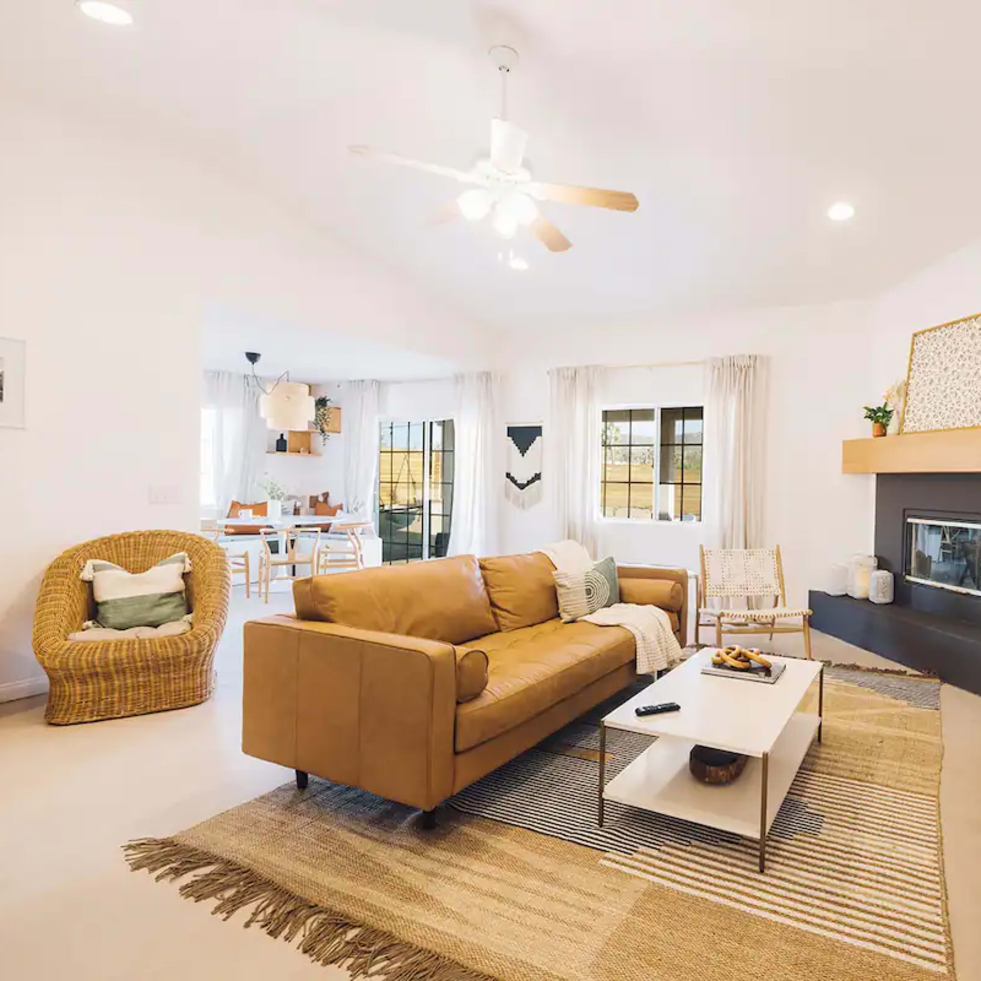 The living room features a tan sofa, a coffee table with snacks, and a wall-mounted television above a modern fireplace, with a dining area visible in the background.