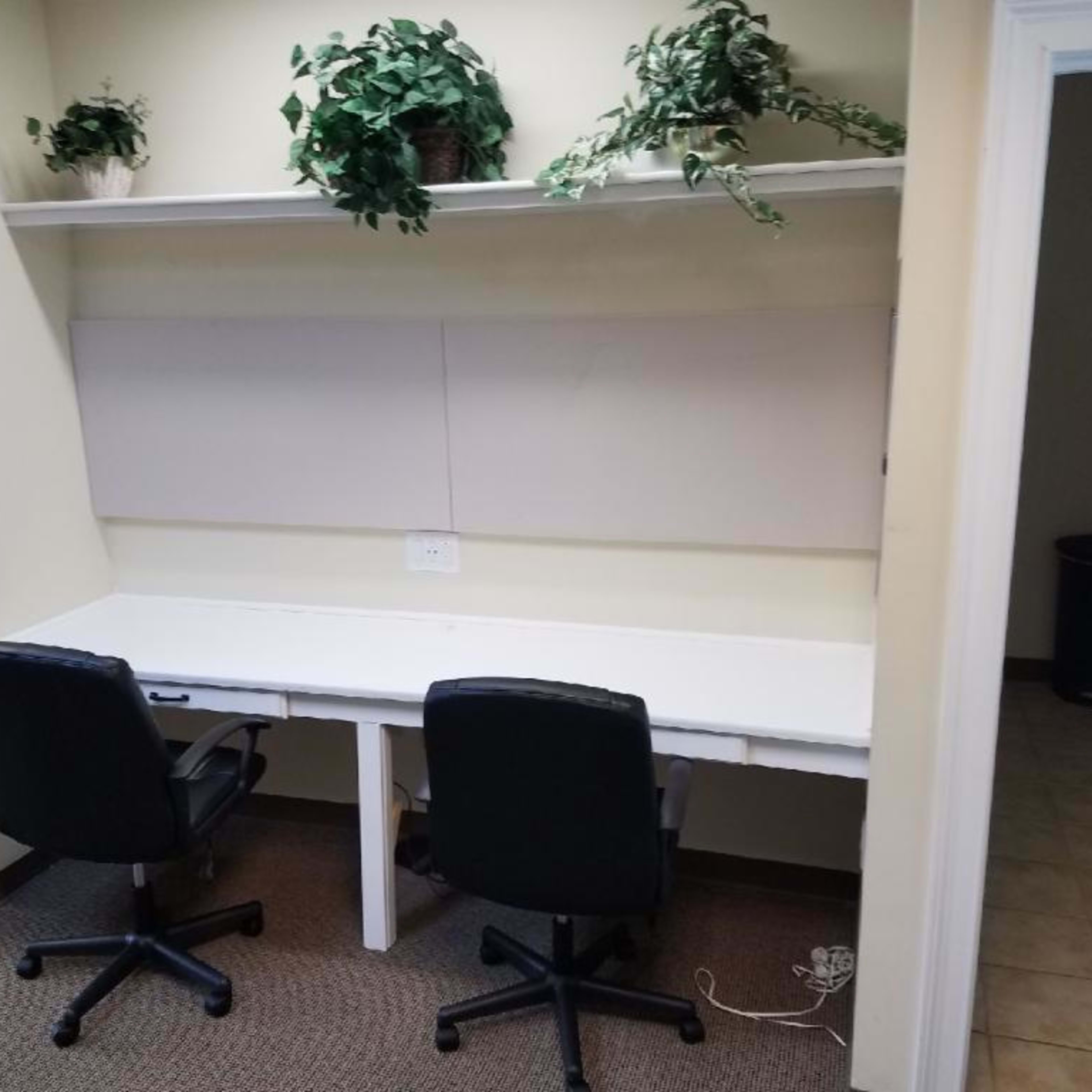 A small office space features two black chairs positioned at a white desk with plants on a shelf above and a door leading to another room on the right.