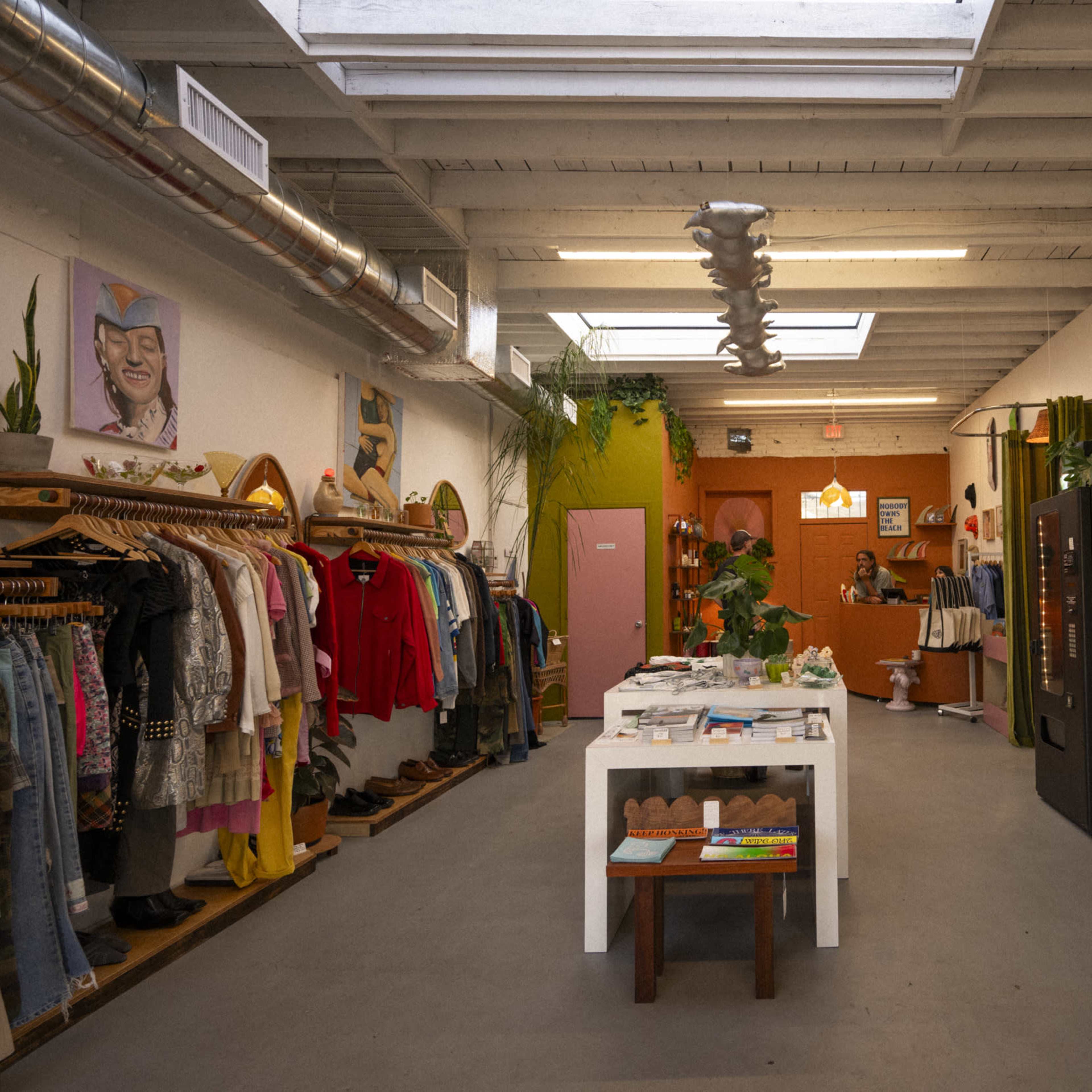 The interior of a boutique features clothing racks on one side, a surfboard display on the other, and a counters area with a cashier at the back.