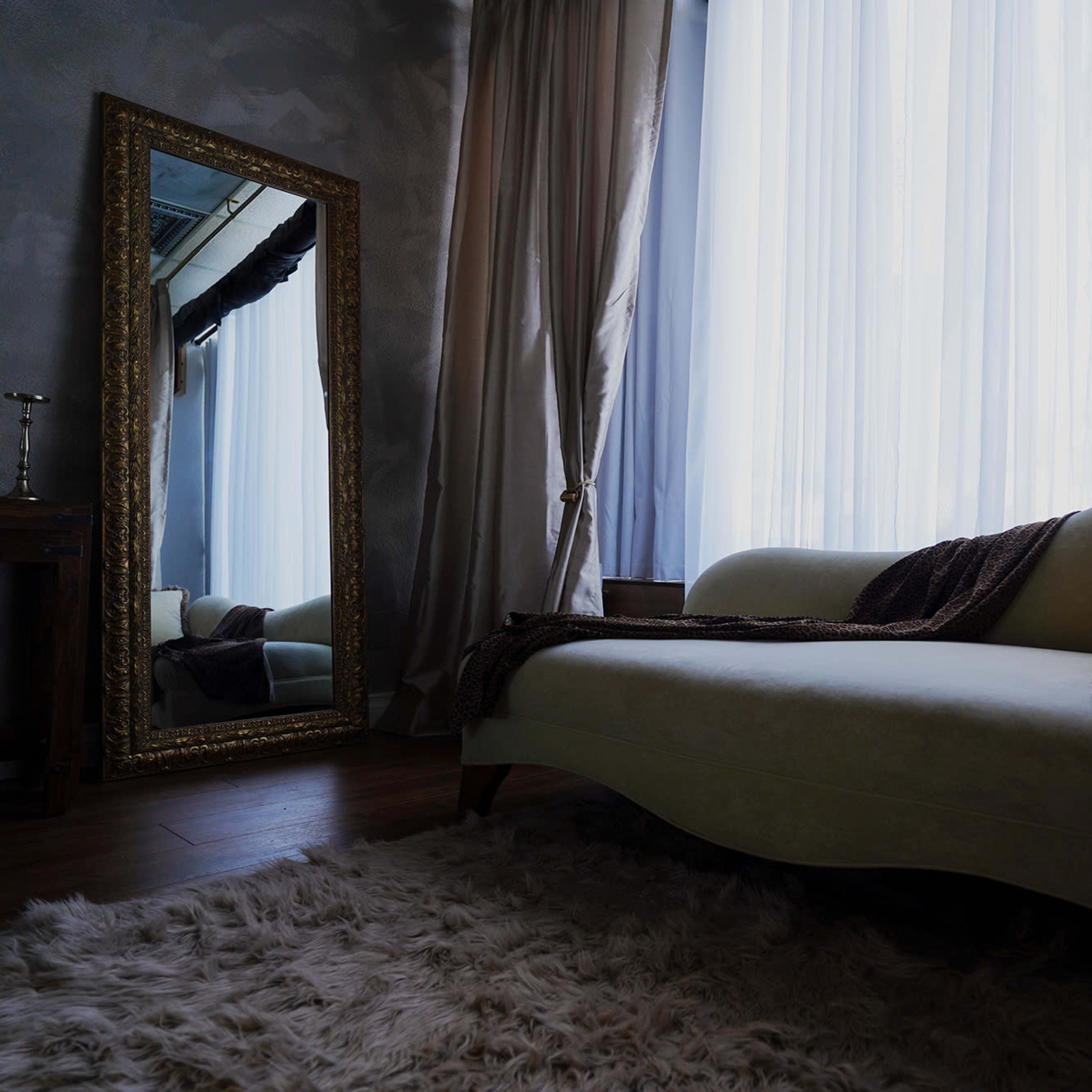 A light green chaise lounge is placed beside a large ornate mirror in a softly lit room with sheer curtains.
