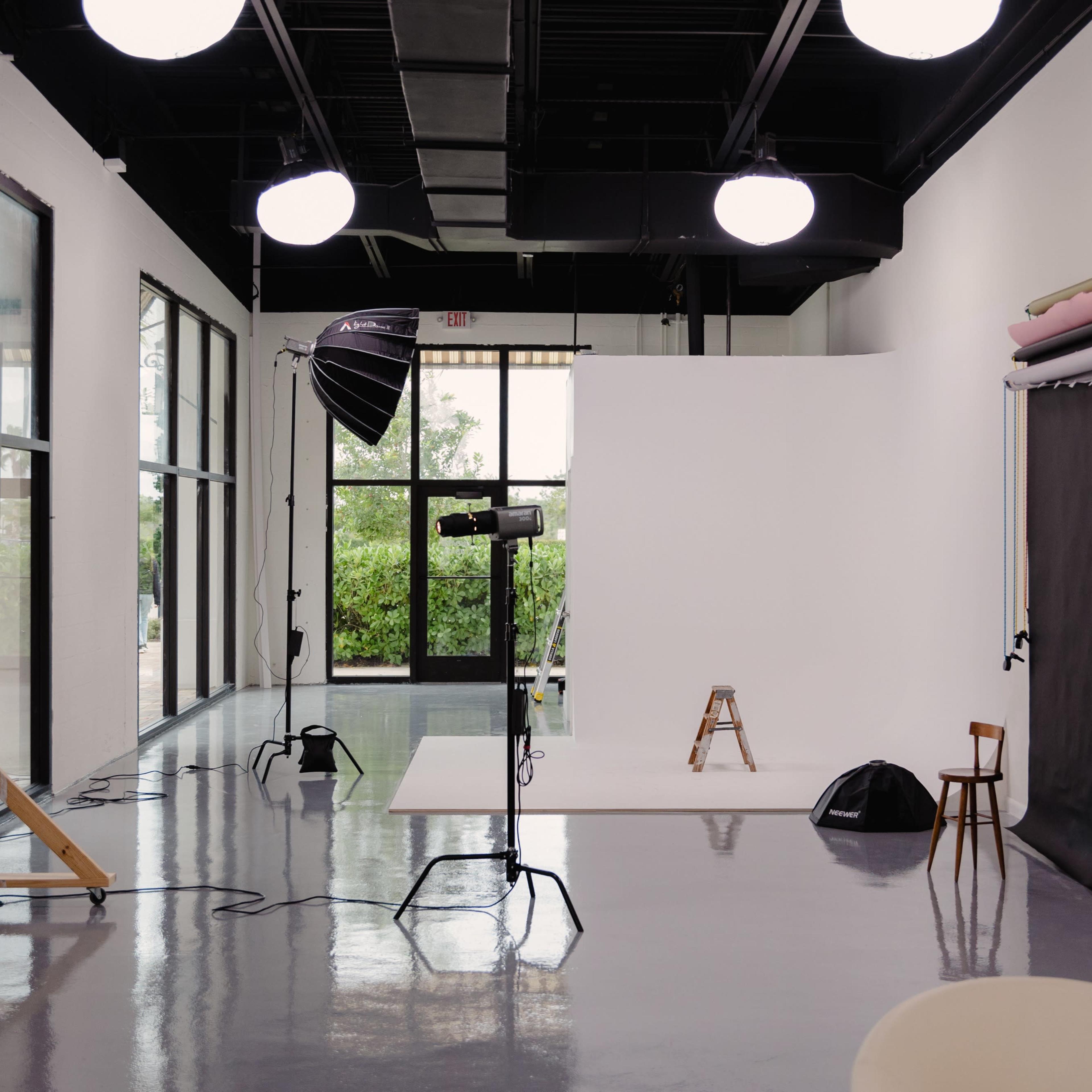 A photography studio with a white backdrop, various lighting equipment, stools, and large windows letting in natural light.
