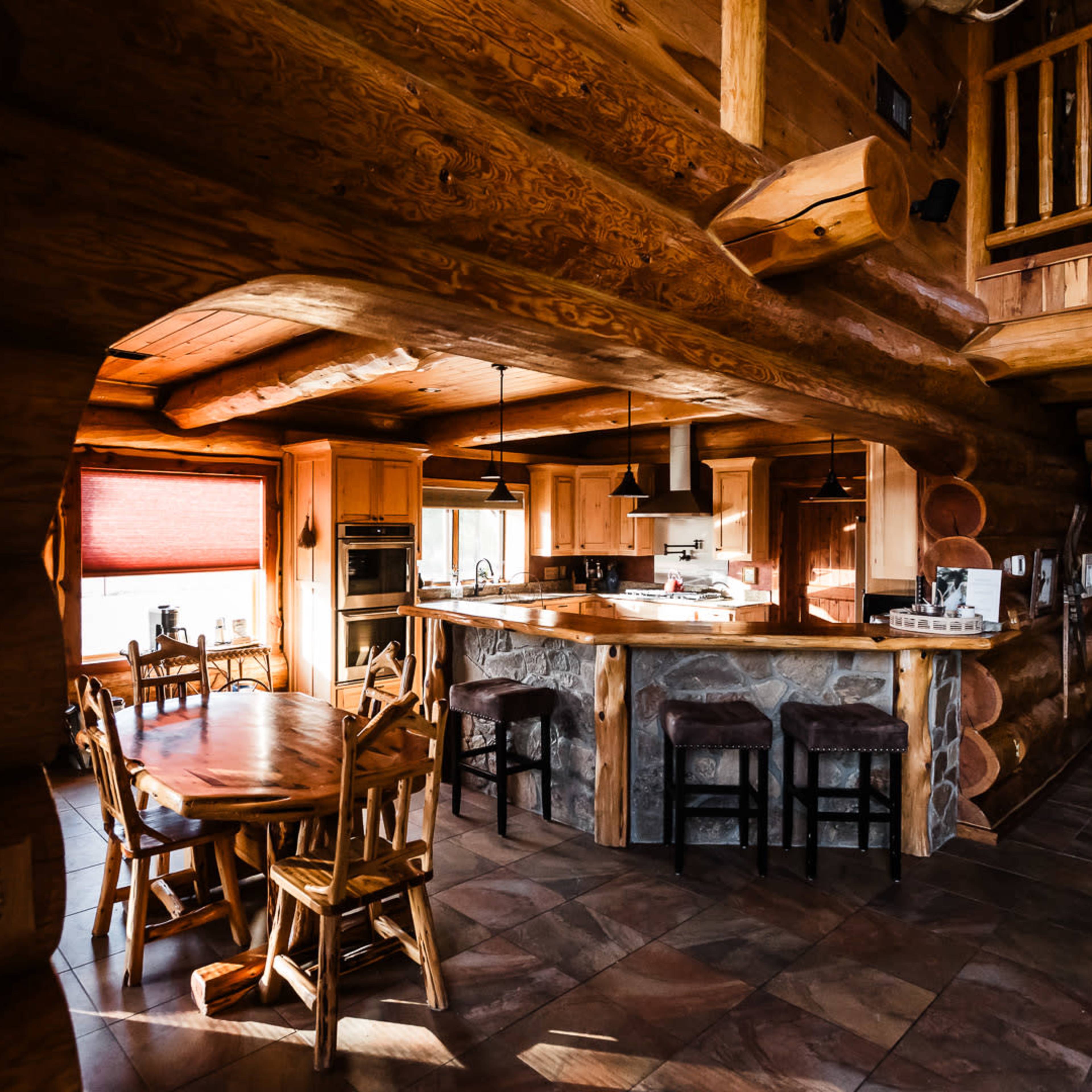 A rustic log cabin interior with a kitchen area, dining table, and wooden furnishings.