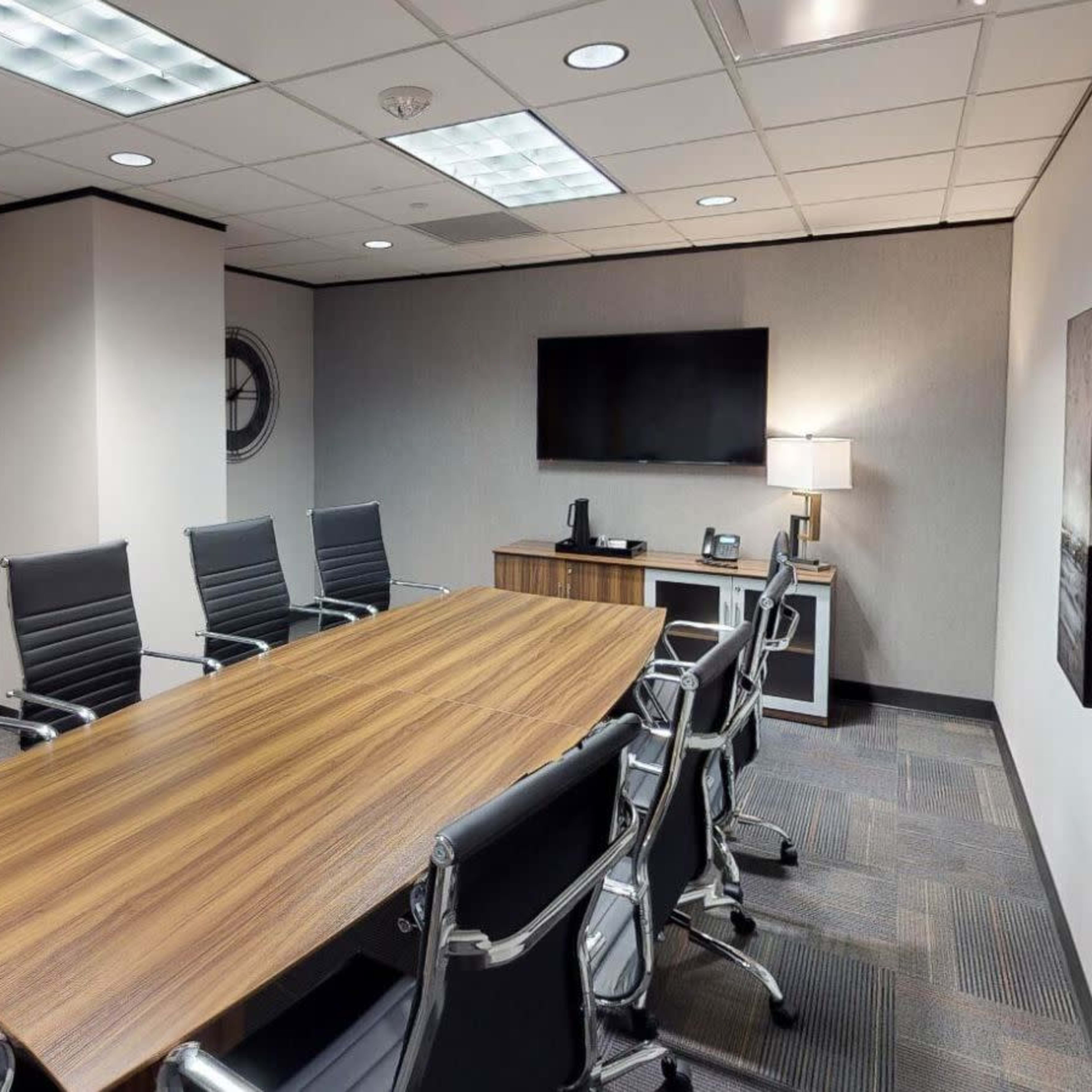 A modern conference room features a long wooden table surrounded by black leather chairs, with a television and a lamp on a side cabinet.