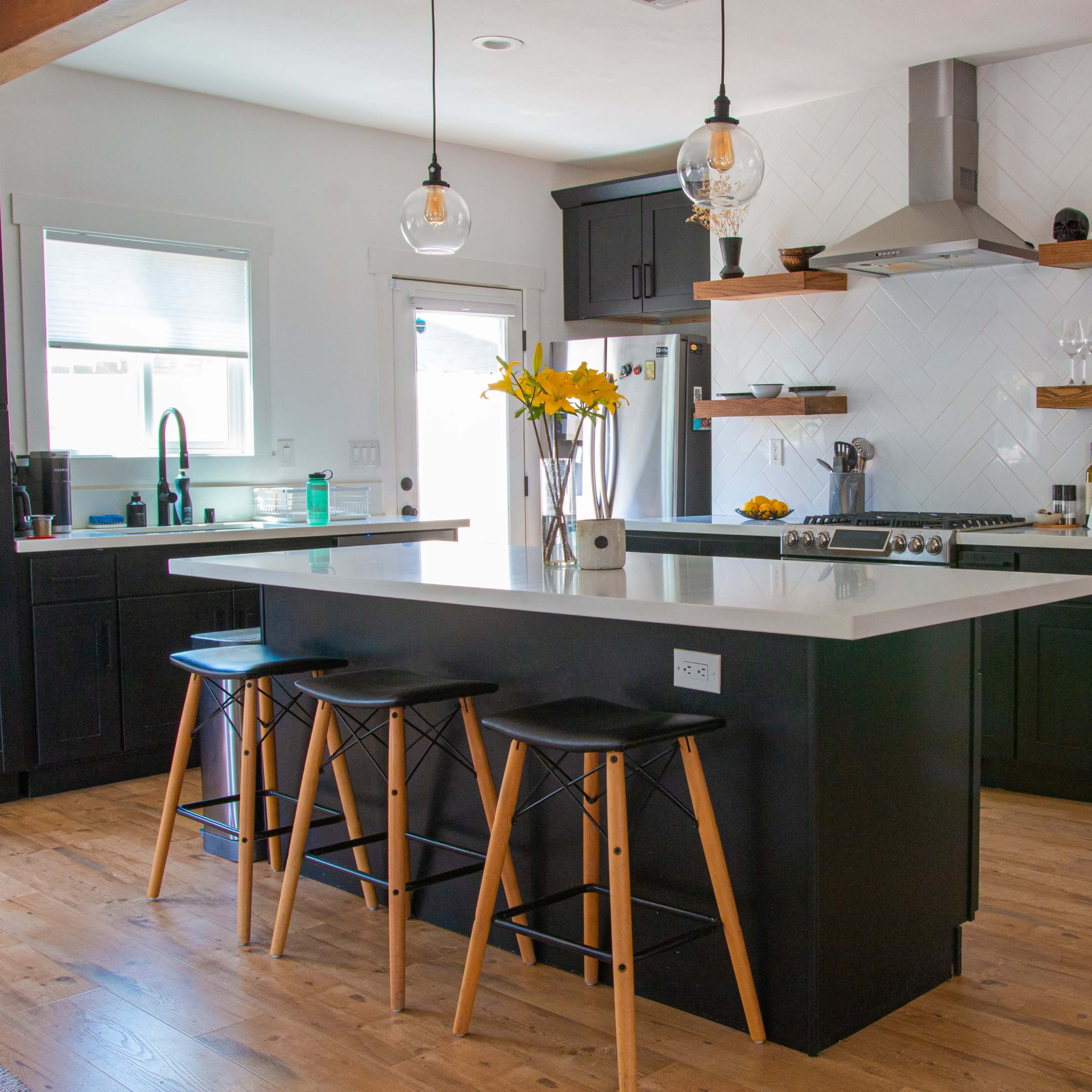 A modern kitchen features a central island with three wooden stools, dark cabinetry, open shelving with decorative items, and a stainless steel range hood above the stove.