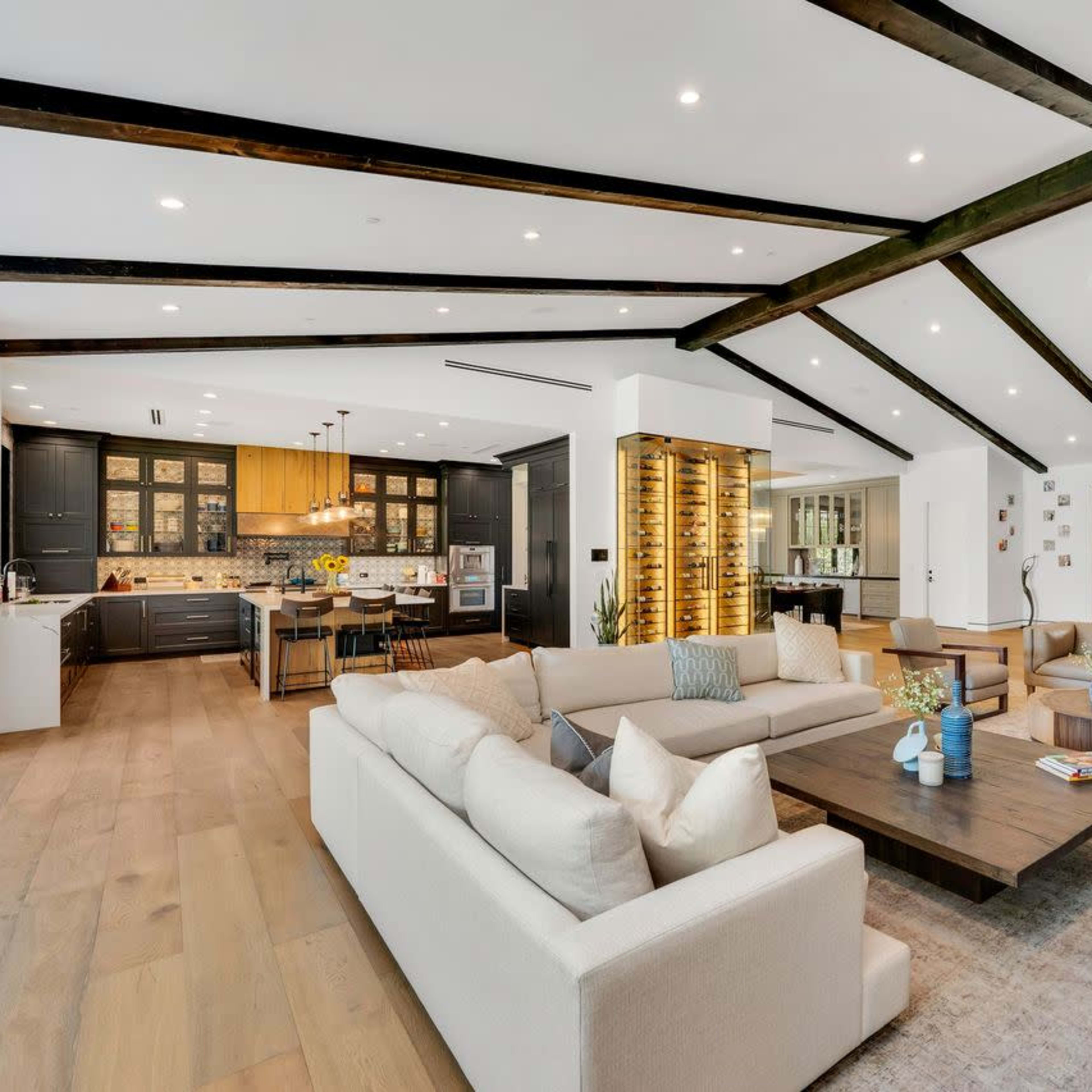 A modern living room with a large sectional sofa, a wooden coffee table, and exposed beam ceilings, opening up to a kitchen area with dark cabinetry.