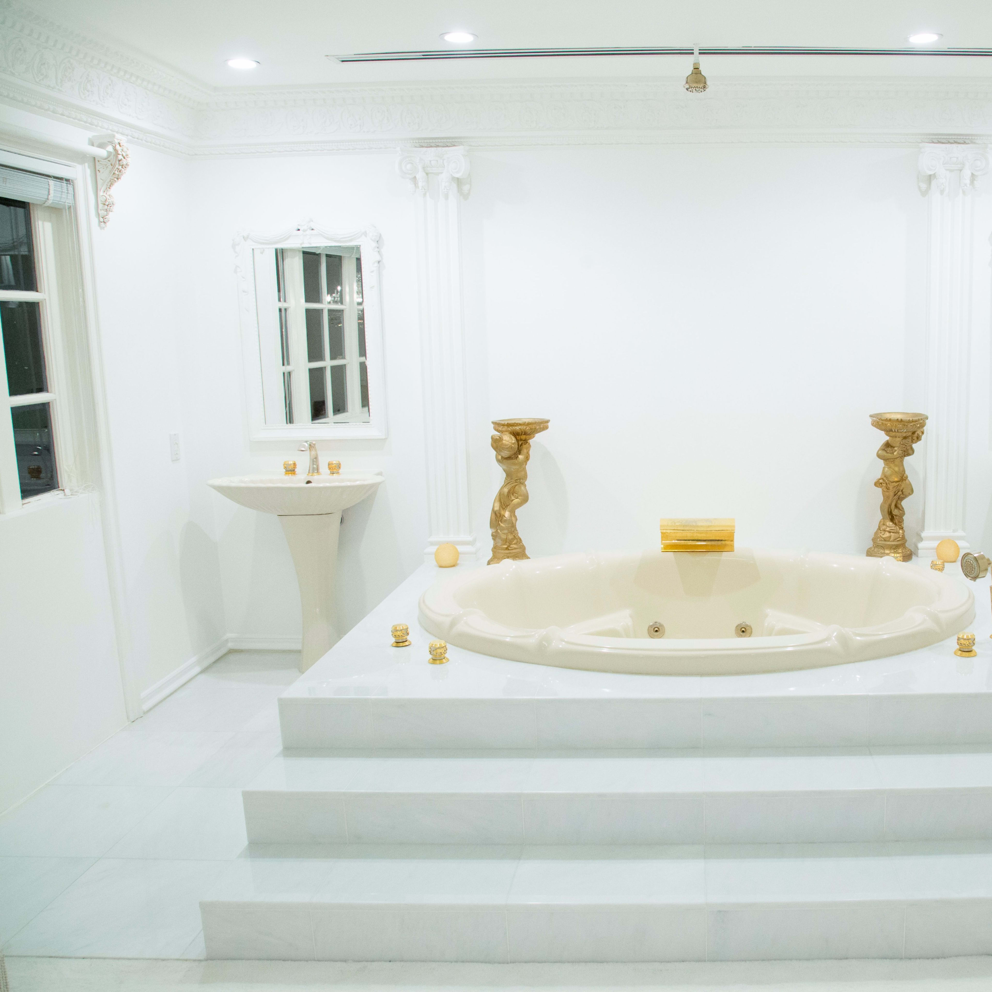 A spacious, brightly lit bathroom with a large oval bathtub, two matching sinks on pedestals, and ornate decor elements.