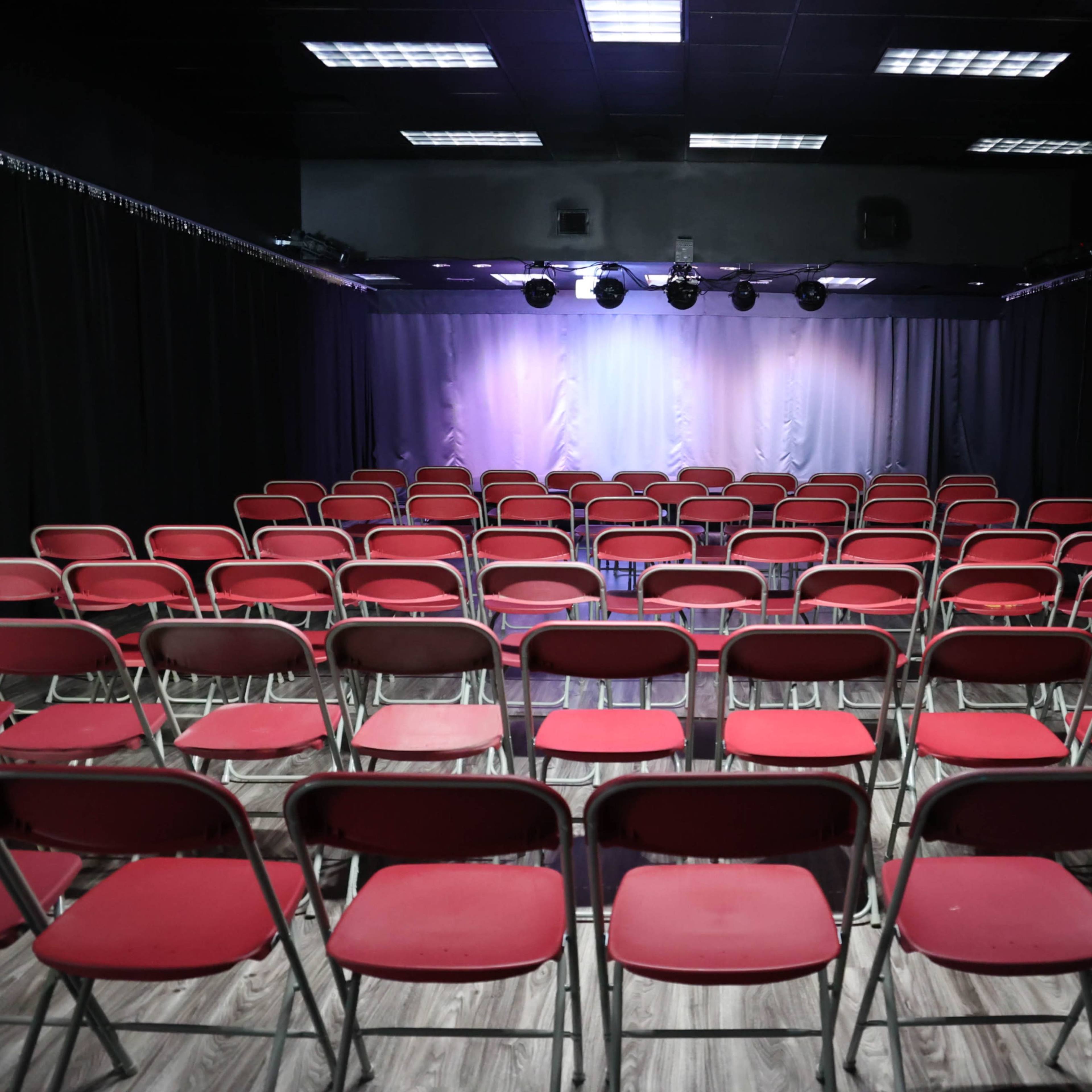 A theater with rows of red chairs facing a stage illuminated by overhead lights.