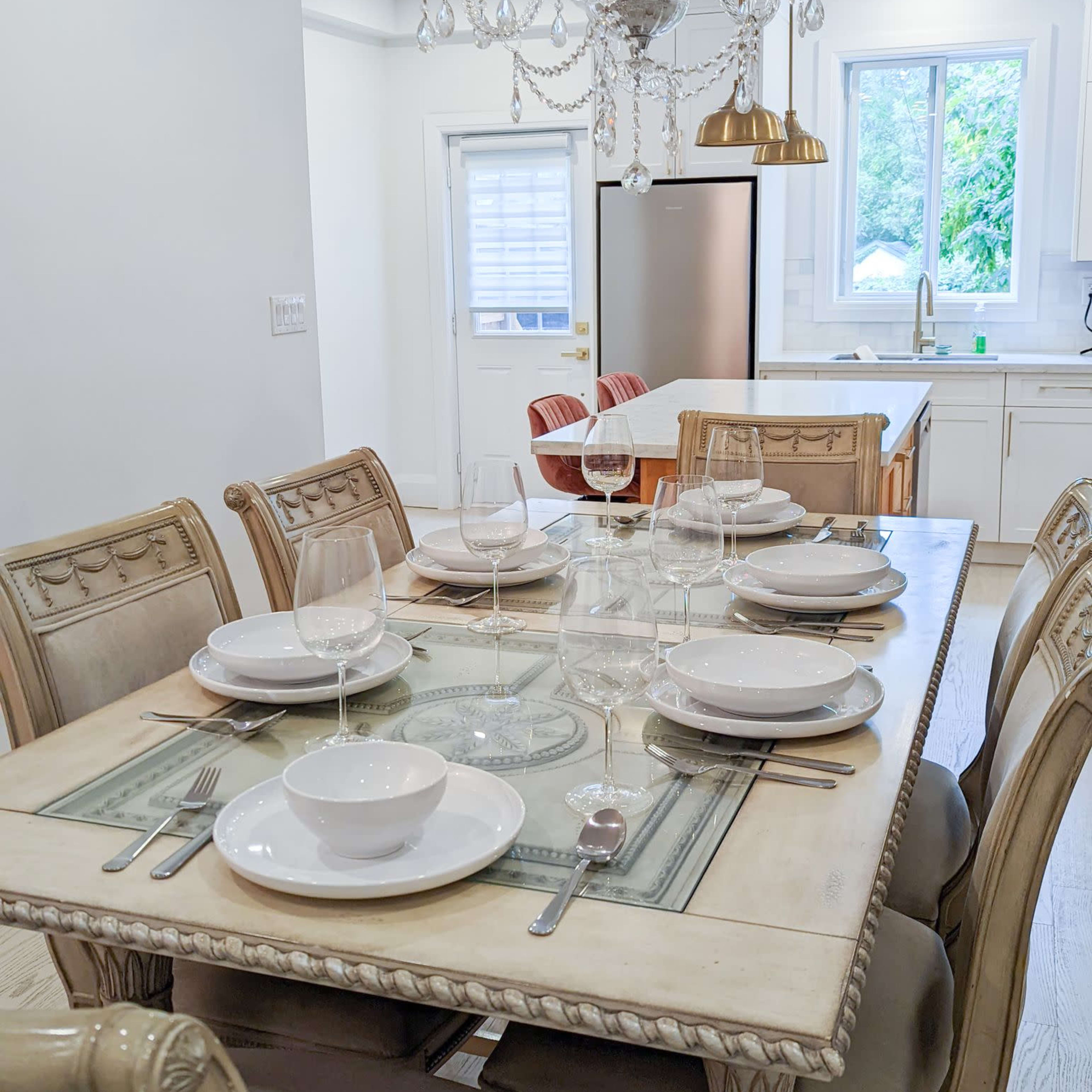 A dining table is set with plates, bowls, and glasses in a well-lit kitchen featuring a chandelier and modern appliances.