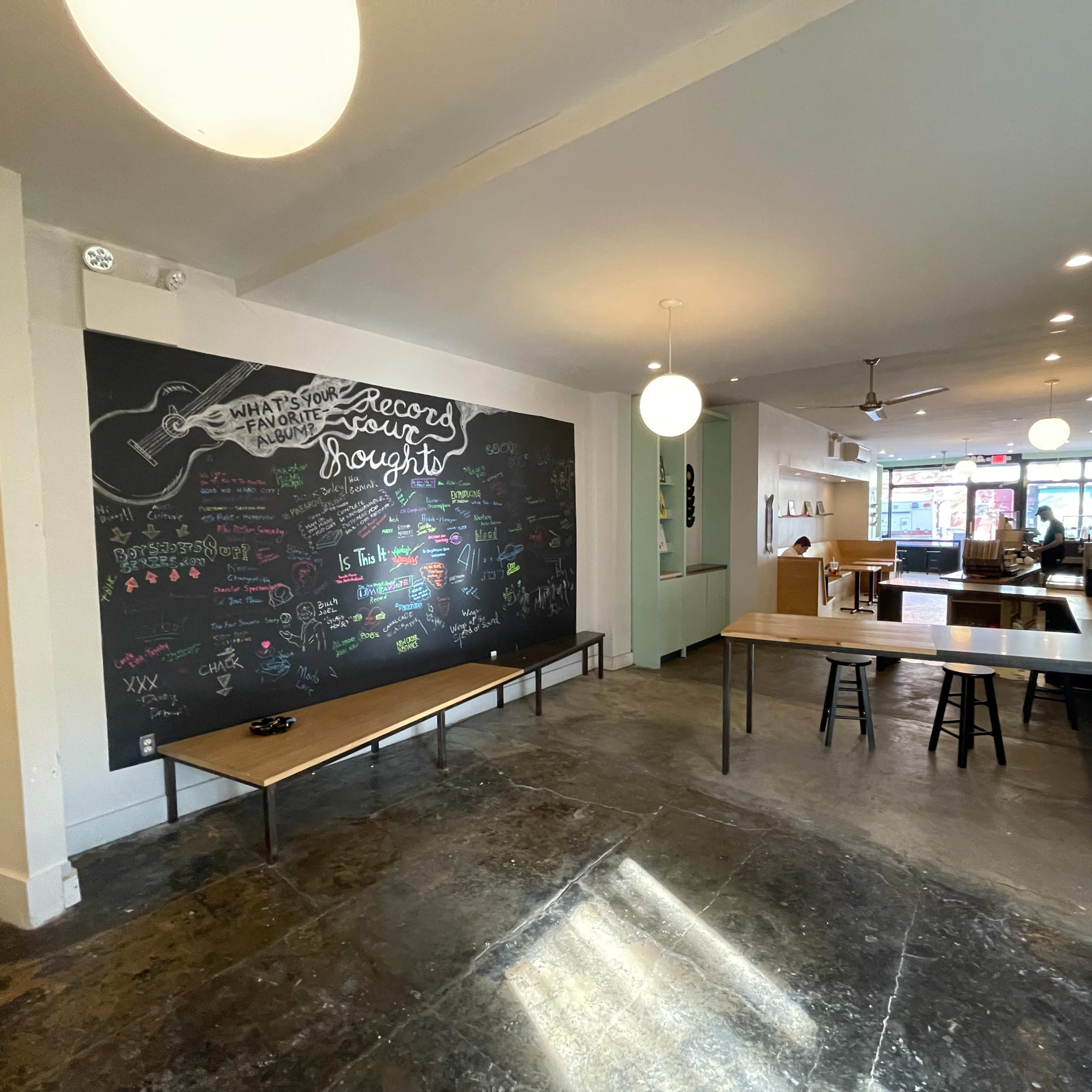 A spacious café interior featuring a large chalkboard wall with colorful writing and two wooden tables set up near the floor.