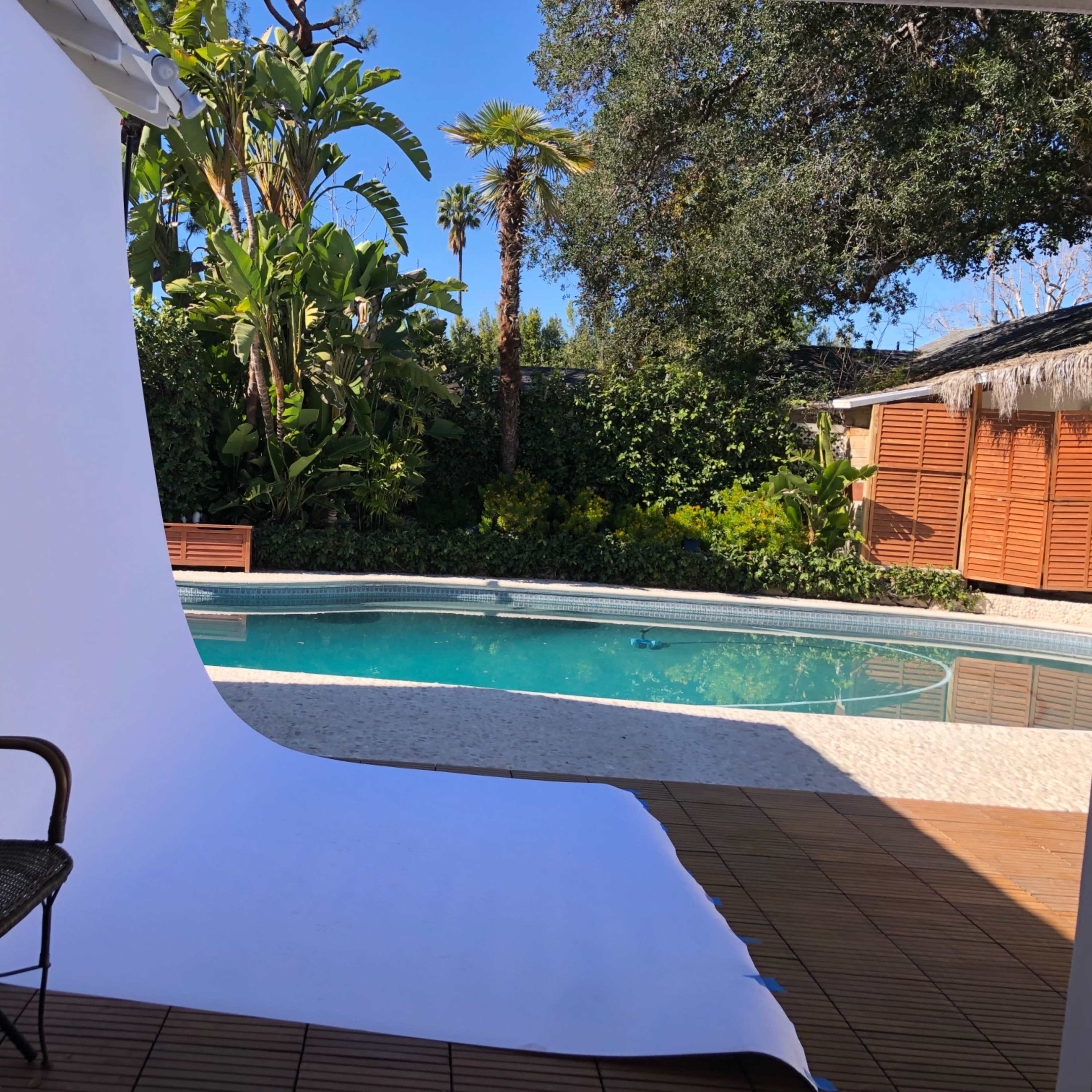 A white backdrop is set up next to a swimming pool surrounded by tropical plants and wooden fencing.