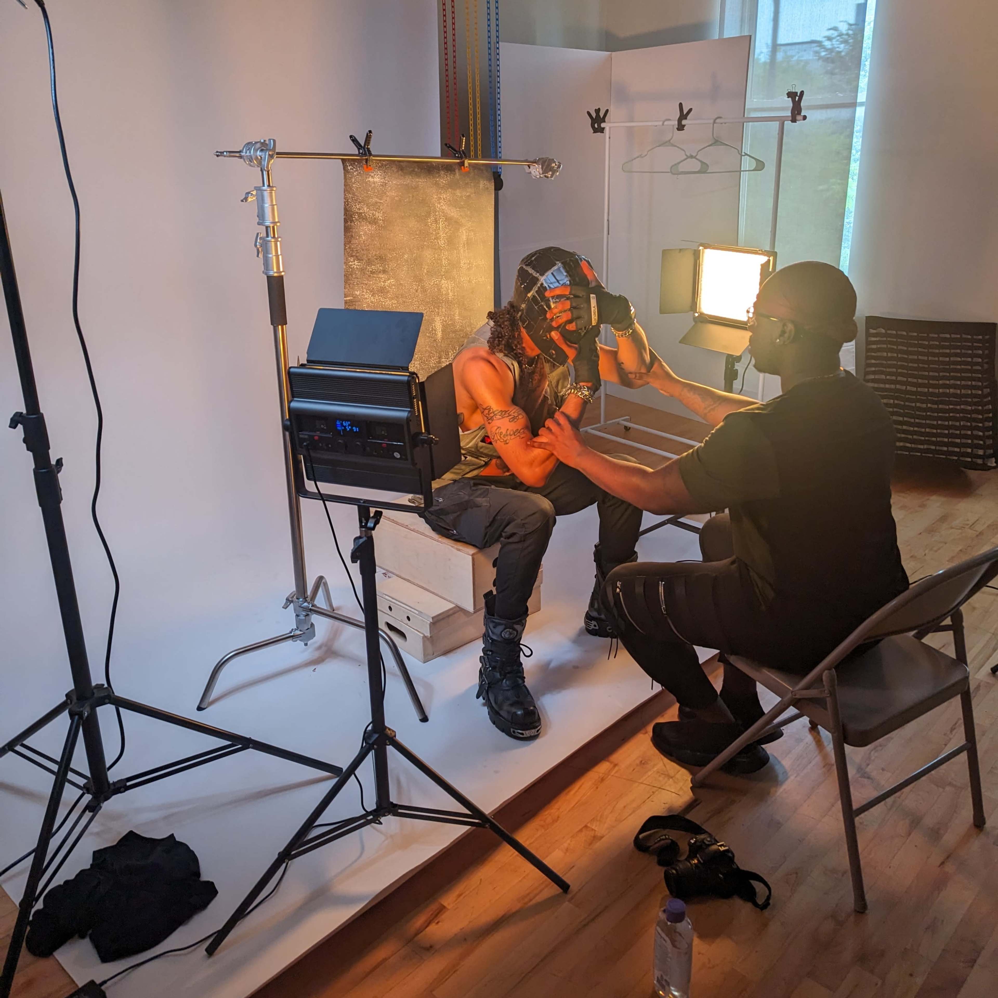 A person with long hair and a patterned head covering sits in front of a lighting setup while another individual adjusts equipment.