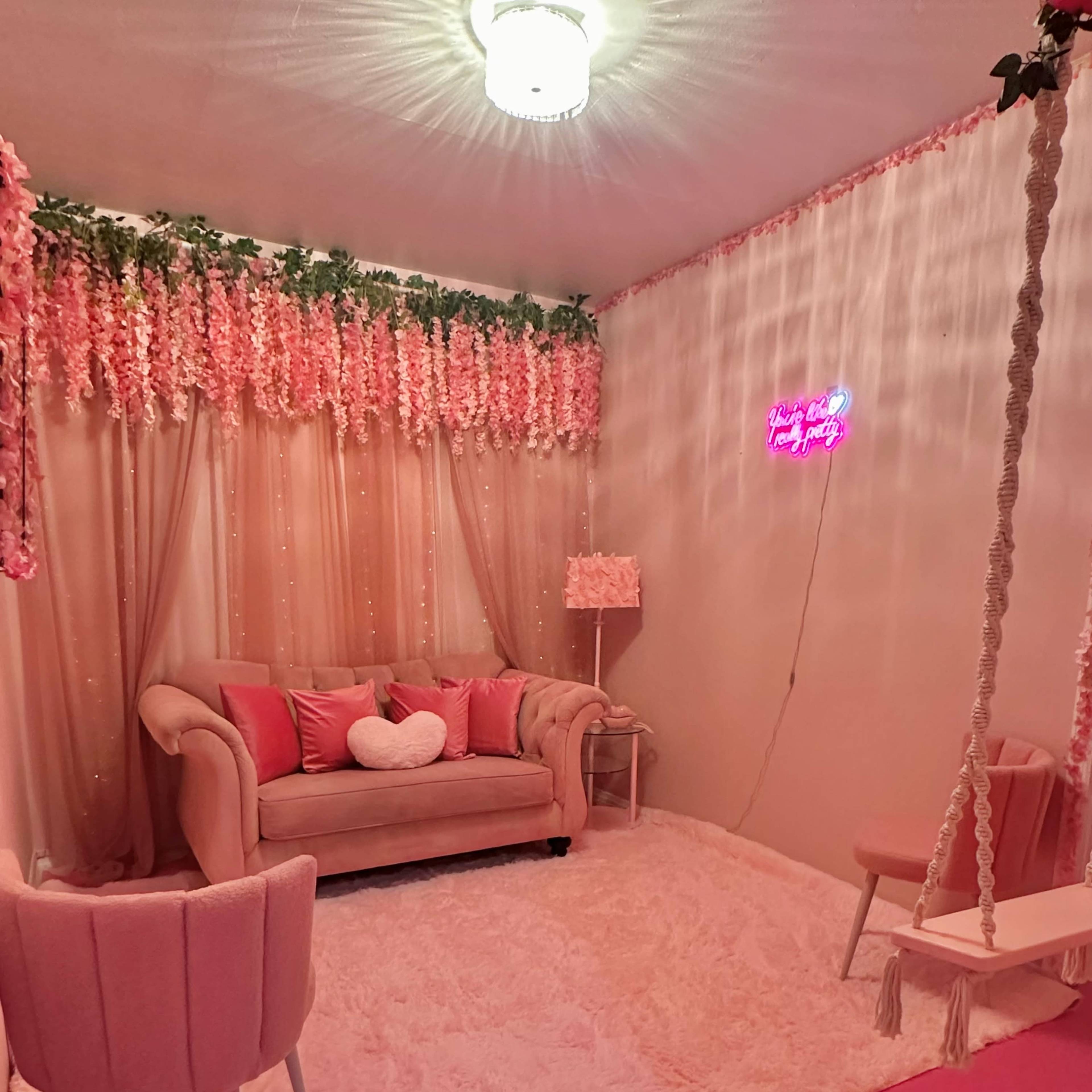A decorated indoor space with a pink-themed sitting area featuring a plush couch, two chairs, and a swing, surrounded by floral arrangements and soft lighting.