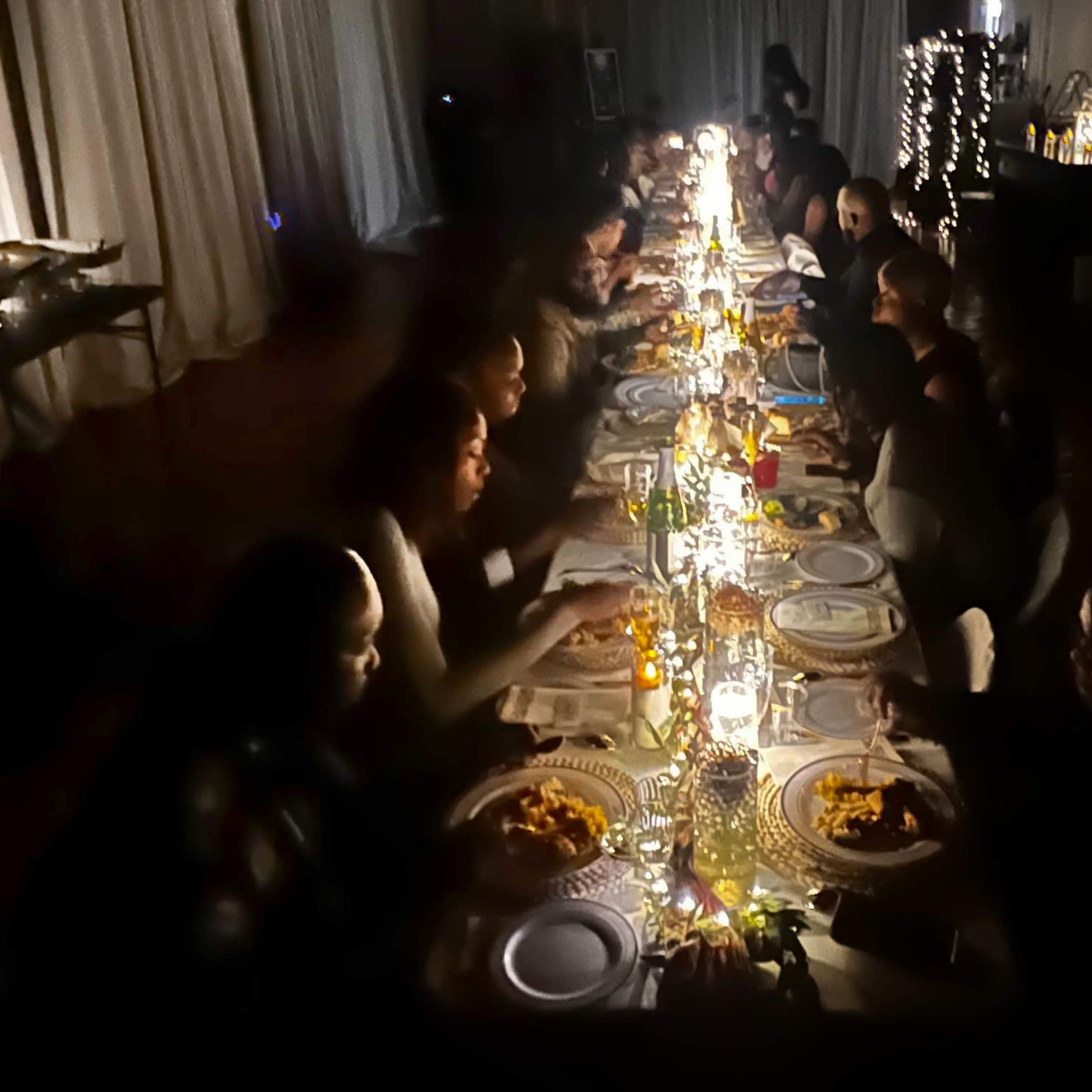 A long dining table is set for a gathering with people eating and socializing under dim, ambient lighting.