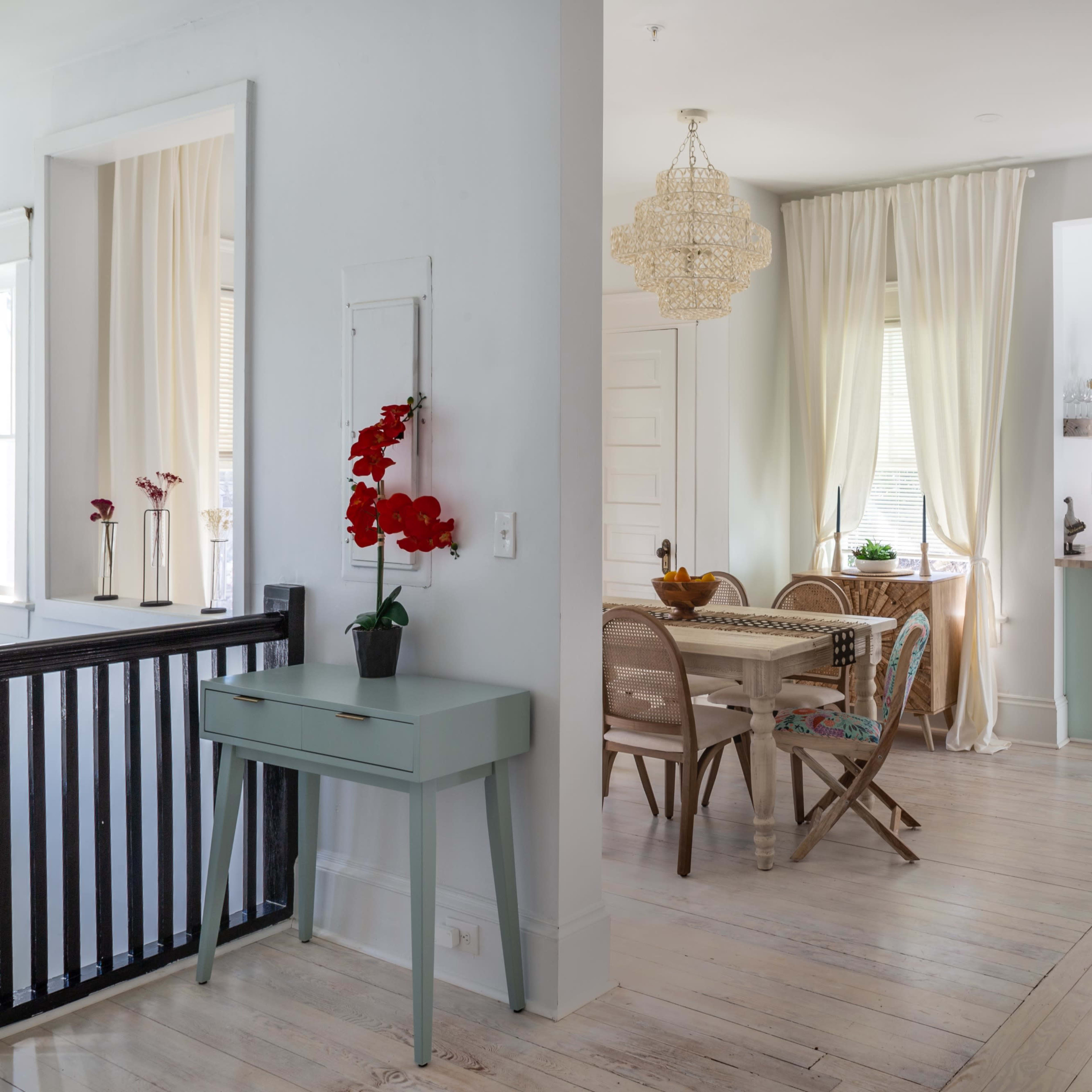 A brightly lit interior features a dining area with a wooden table and chairs, adjoining a modern kitchen with mint green cabinets and a large window.