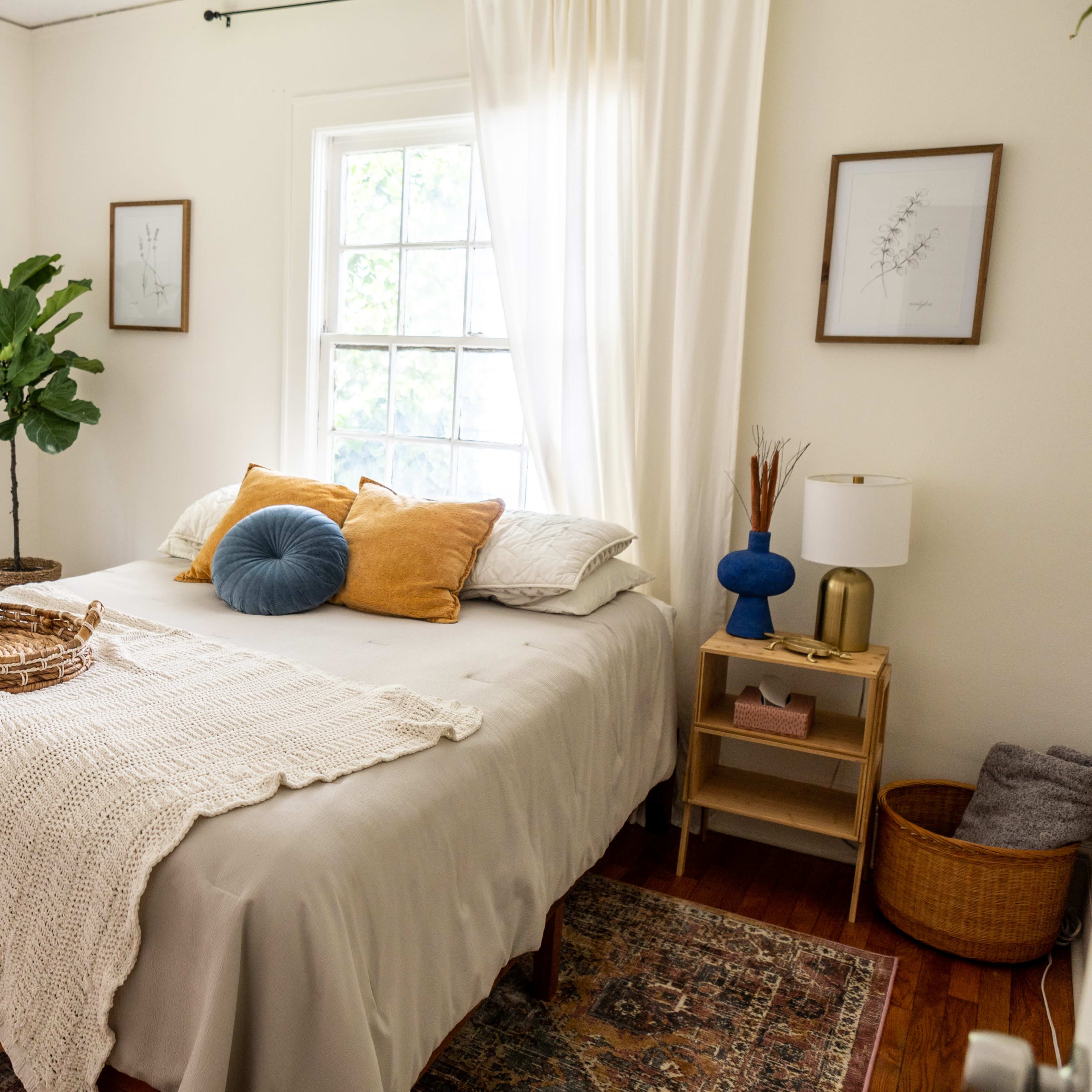 A cozy bedroom features a bed with decorative pillows, a textured throw, and a small bedside table beside large windows.
