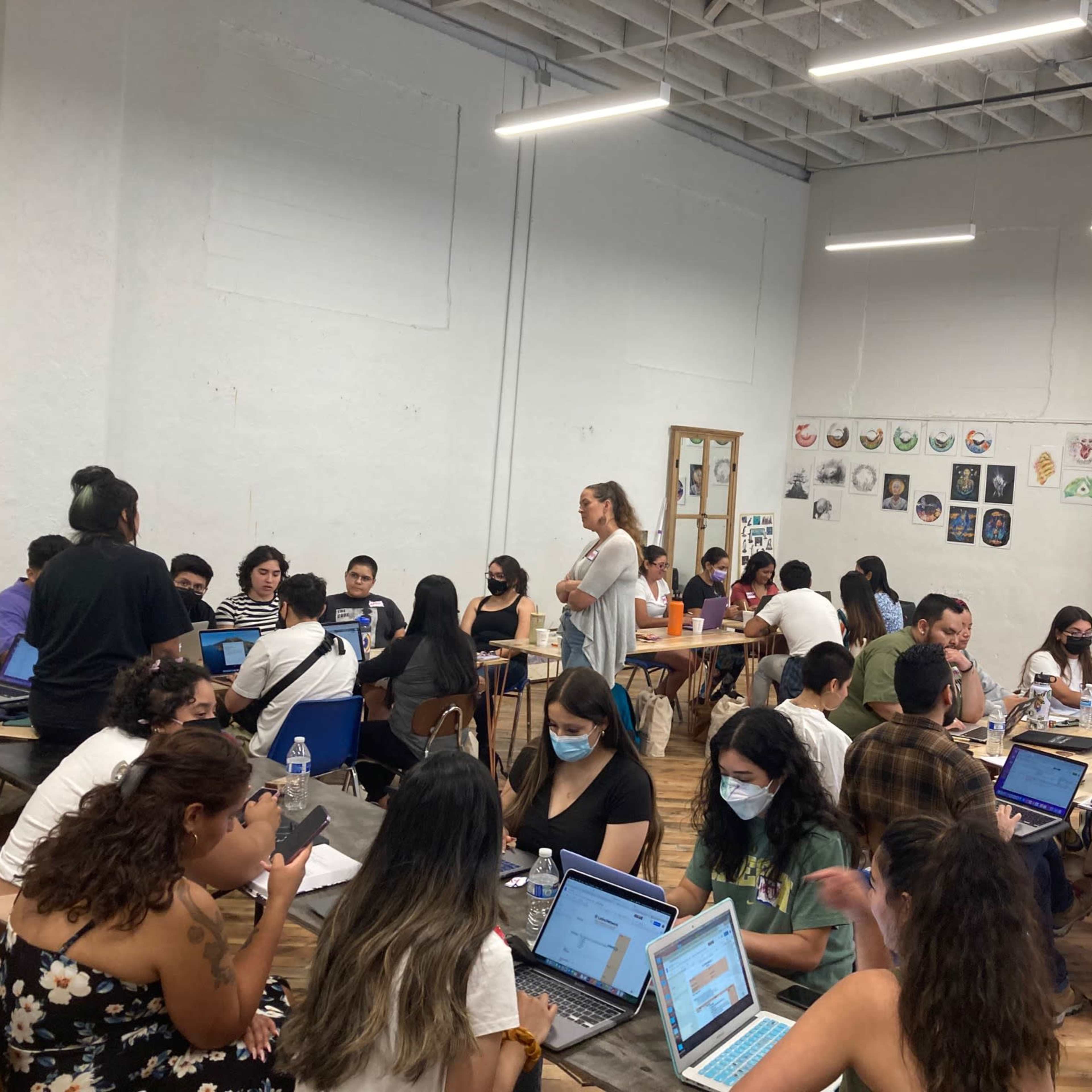 A diverse group of people is engaged in collaborative work at tables in a well-lit room with various artworks displayed on the walls.