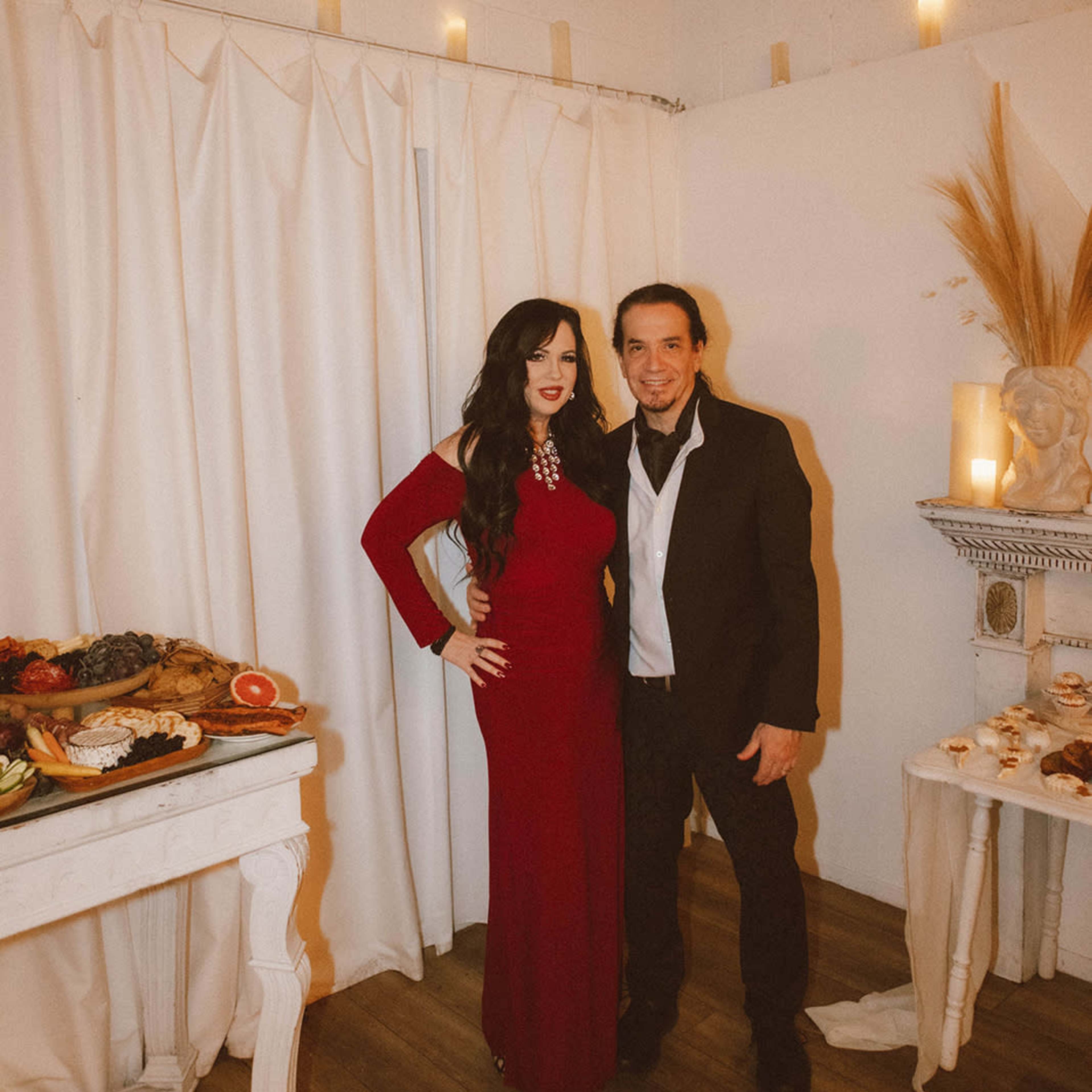 A woman in a red dress and a man in a black suit pose together in front of a food spread and white curtains.
