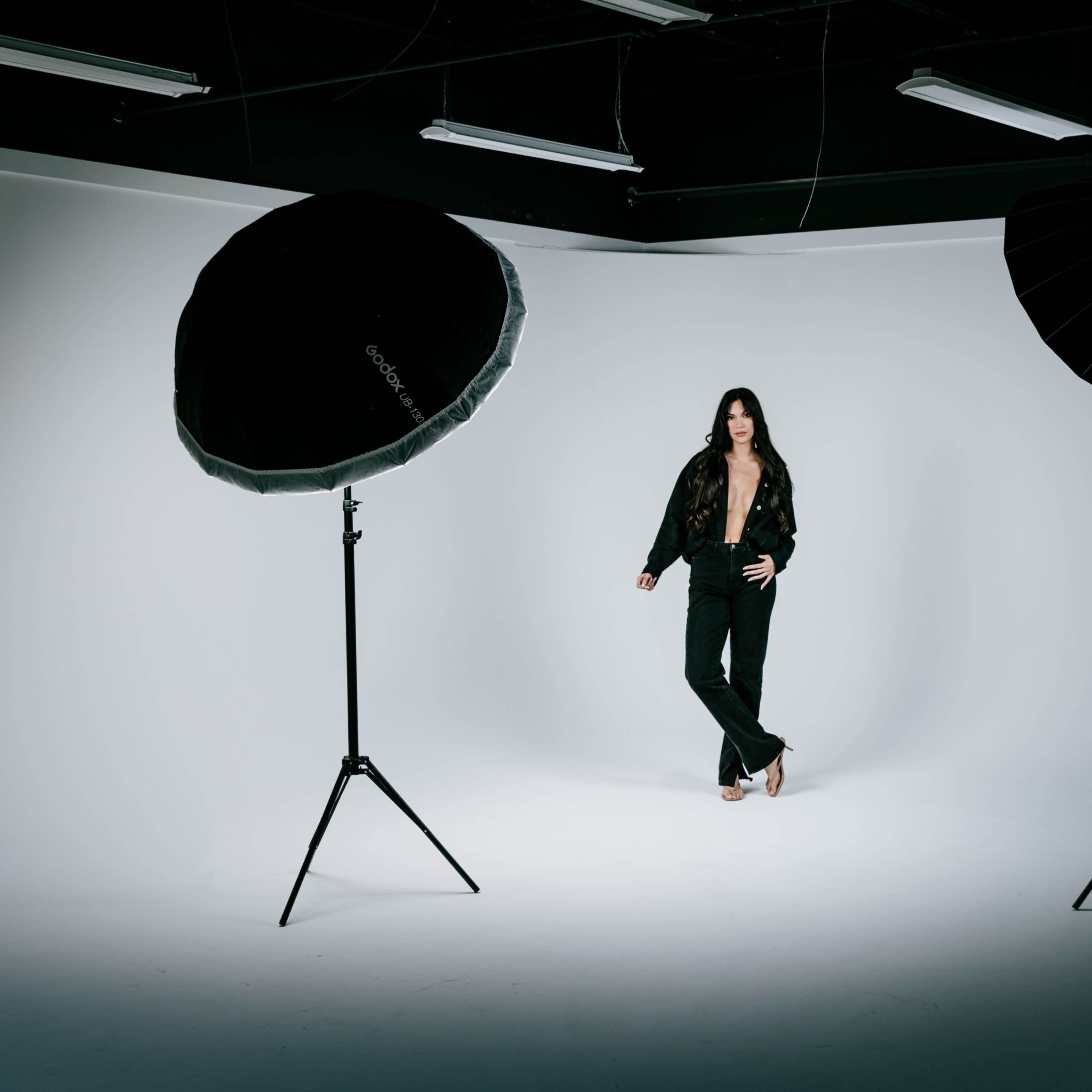 A model poses in a studio surrounded by two large softbox lights against a plain white background.