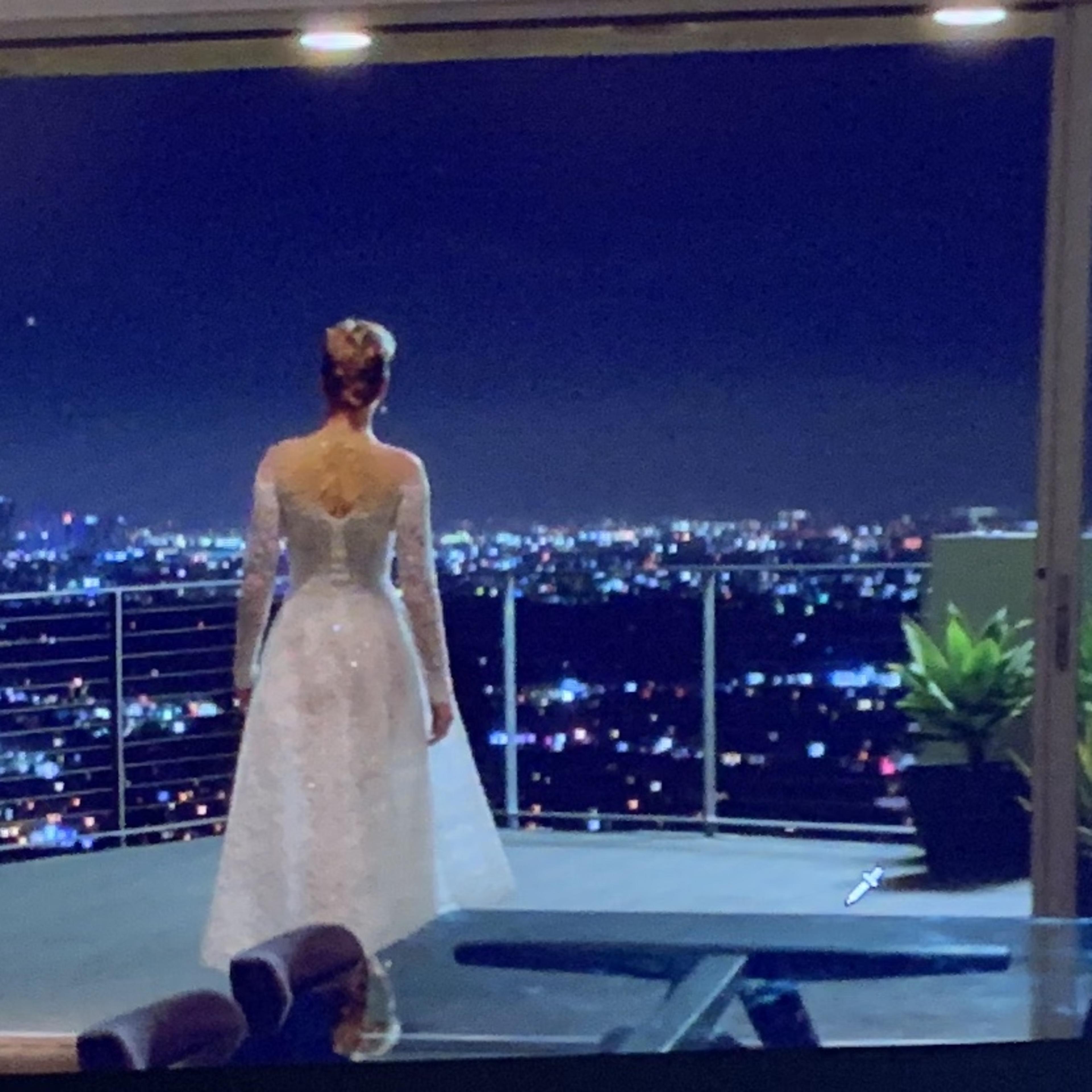 A woman in a white dress stands on a balcony overlooking a city skyline illuminated at night.