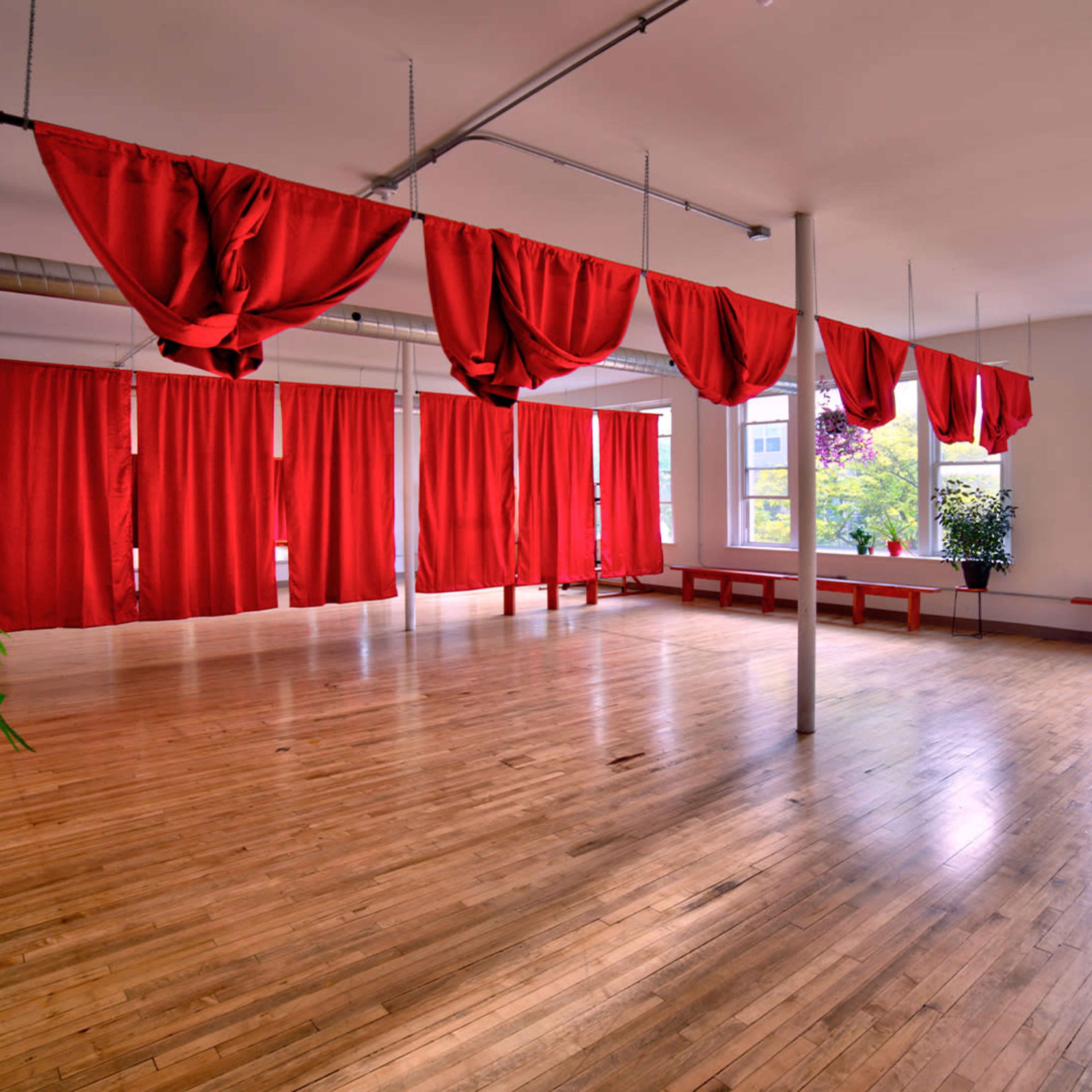A spacious room with wooden flooring, featuring red draped fabric and potted plants near the windows.