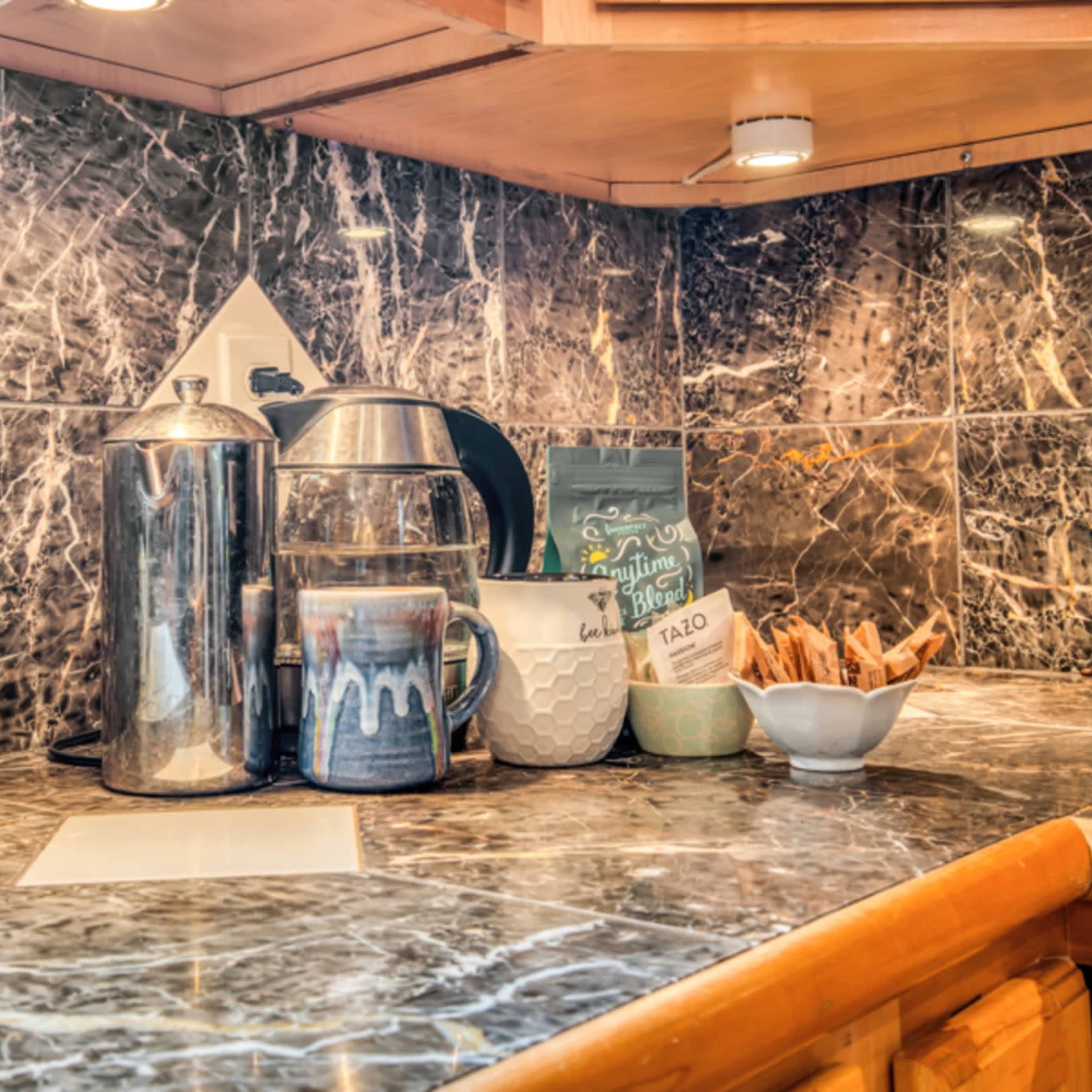 A countertop features a coffee maker, a French press, and various mugs on a marbled surface with wooden cabinetry.