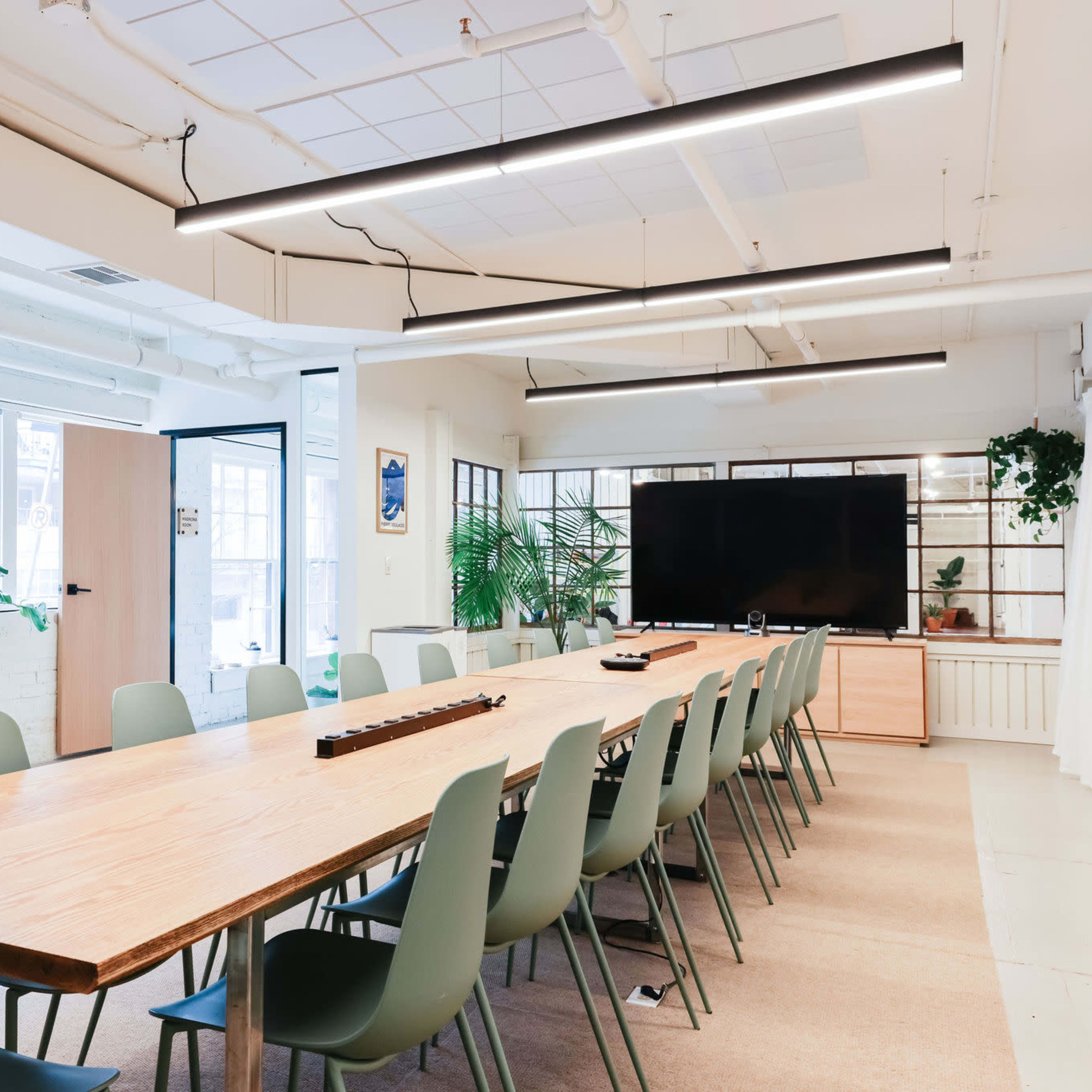 A long wooden table surrounded by green chairs is set up in a bright conference room featuring large windows and a television screen on a wooden stand.