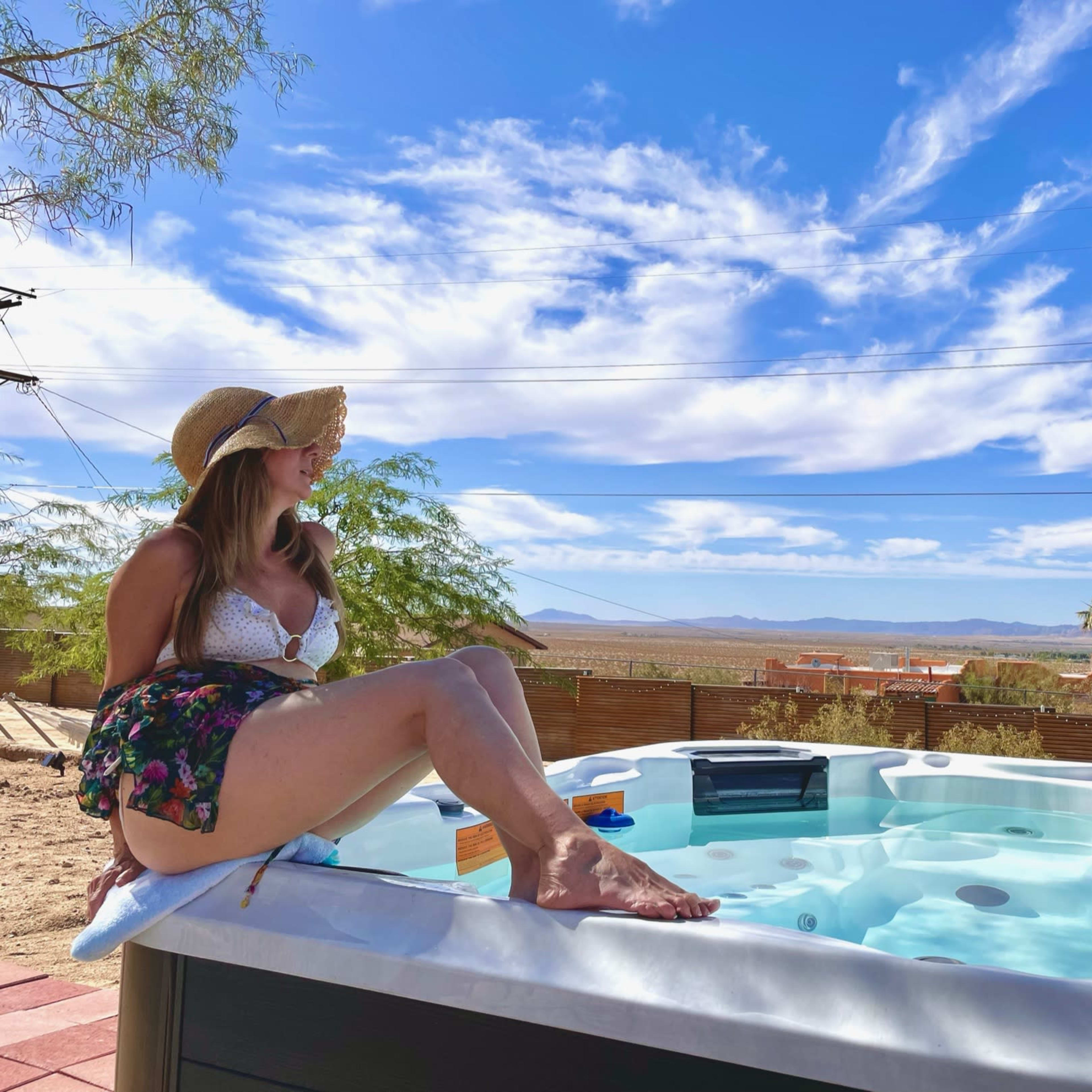 A person in a hat relaxes by a hot tub with a desert landscape and blue sky in the background.