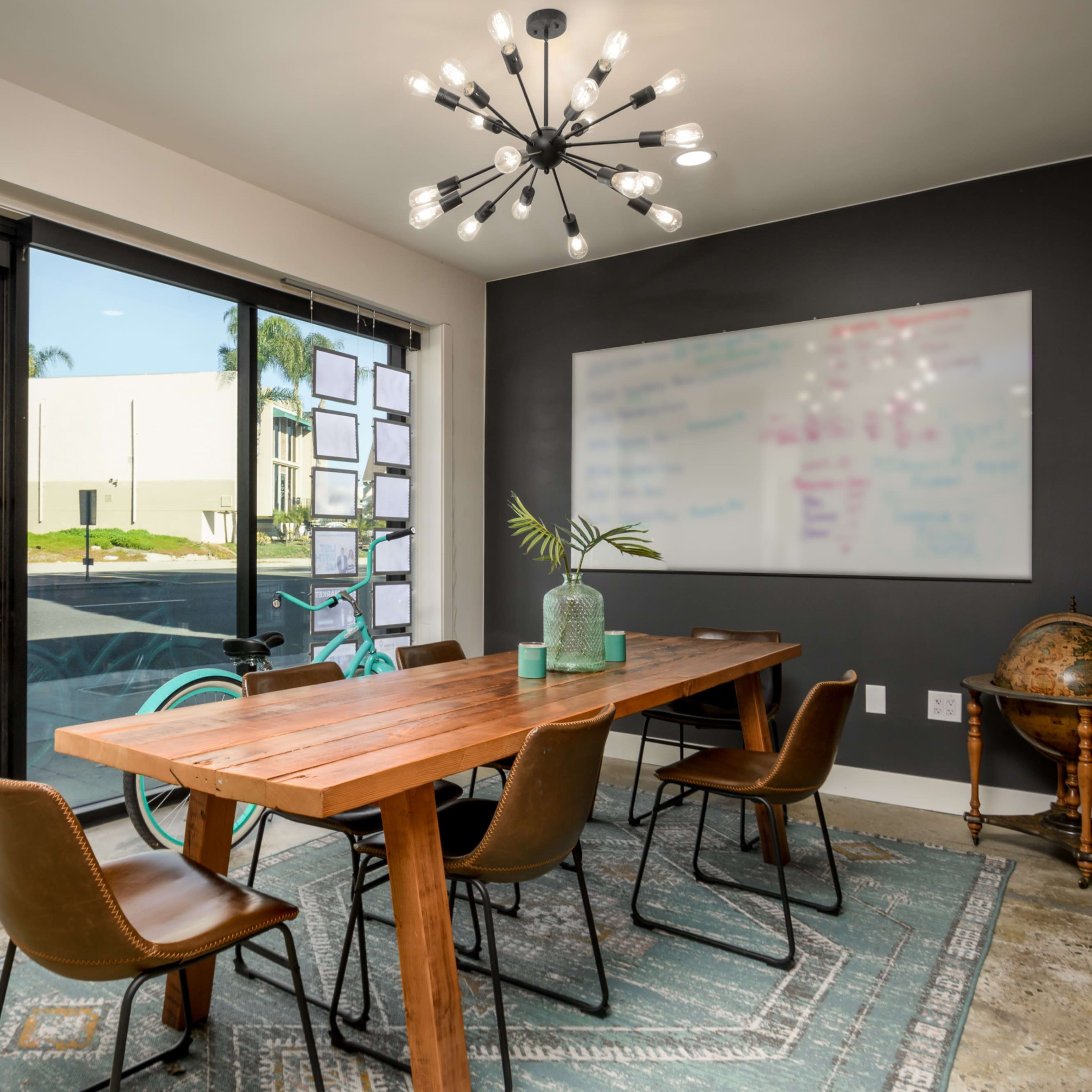 A modern conference room features a wooden table surrounded by leather chairs, large glass doors opening to a view of the street, and a wall-mounted whiteboard.