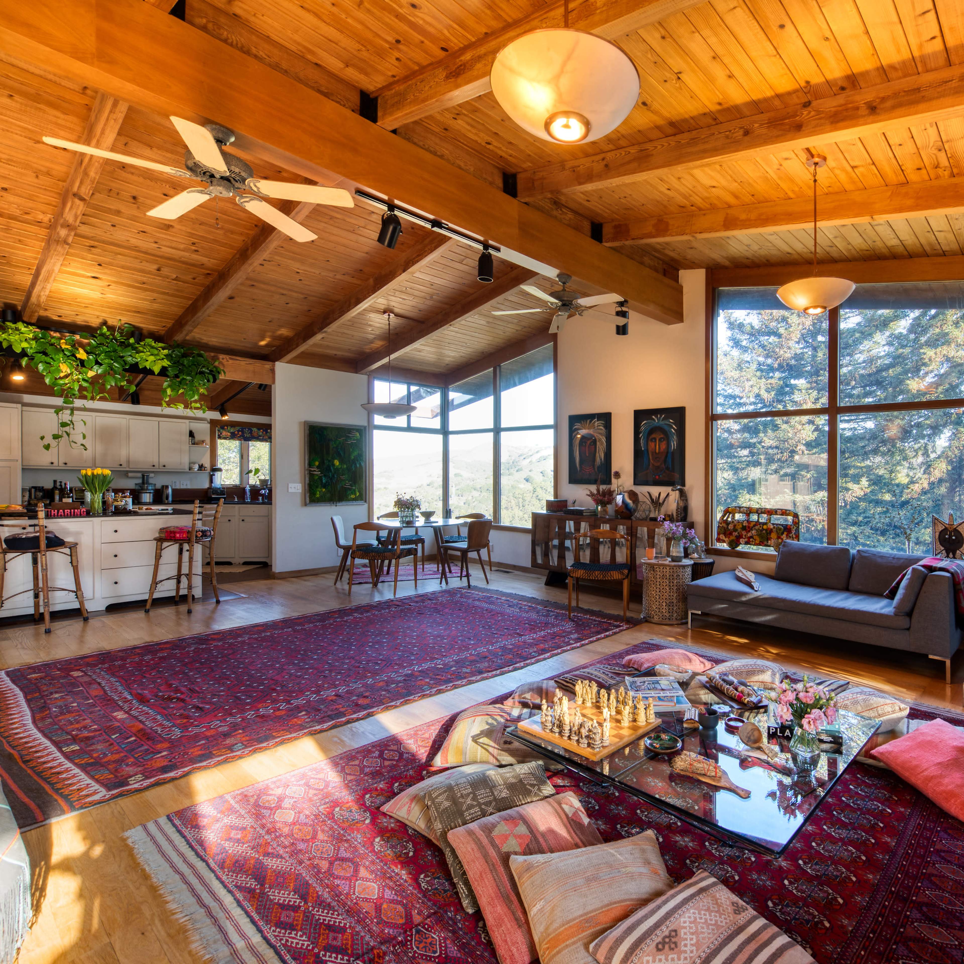 A spacious living area featuring a high wooden ceiling, large windows, a kitchen area, comfortable seating, and various decorative plants.