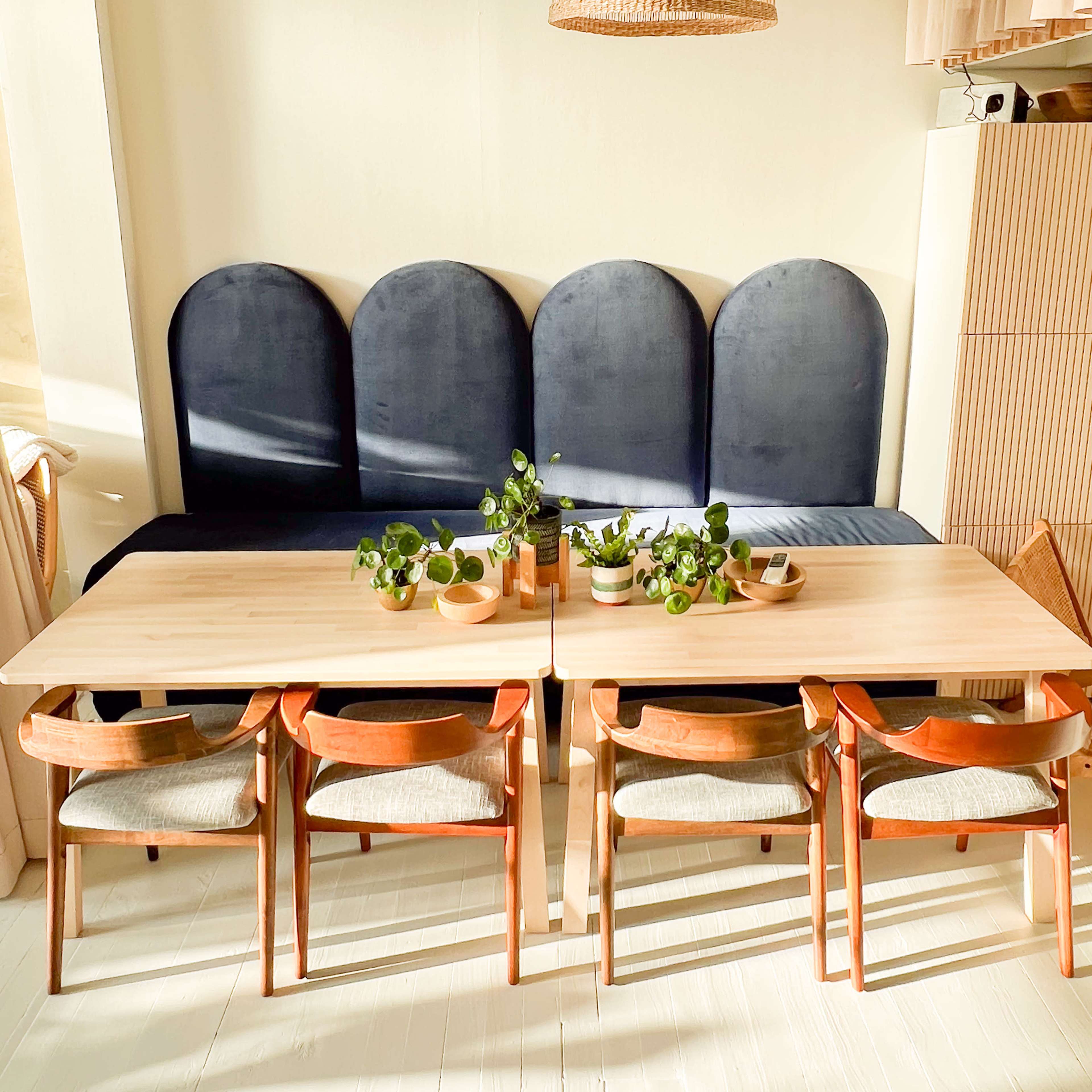 A brightly lit dining area features two wooden tables set with plants, surrounded by chairs facing a cushioned bench along the wall.