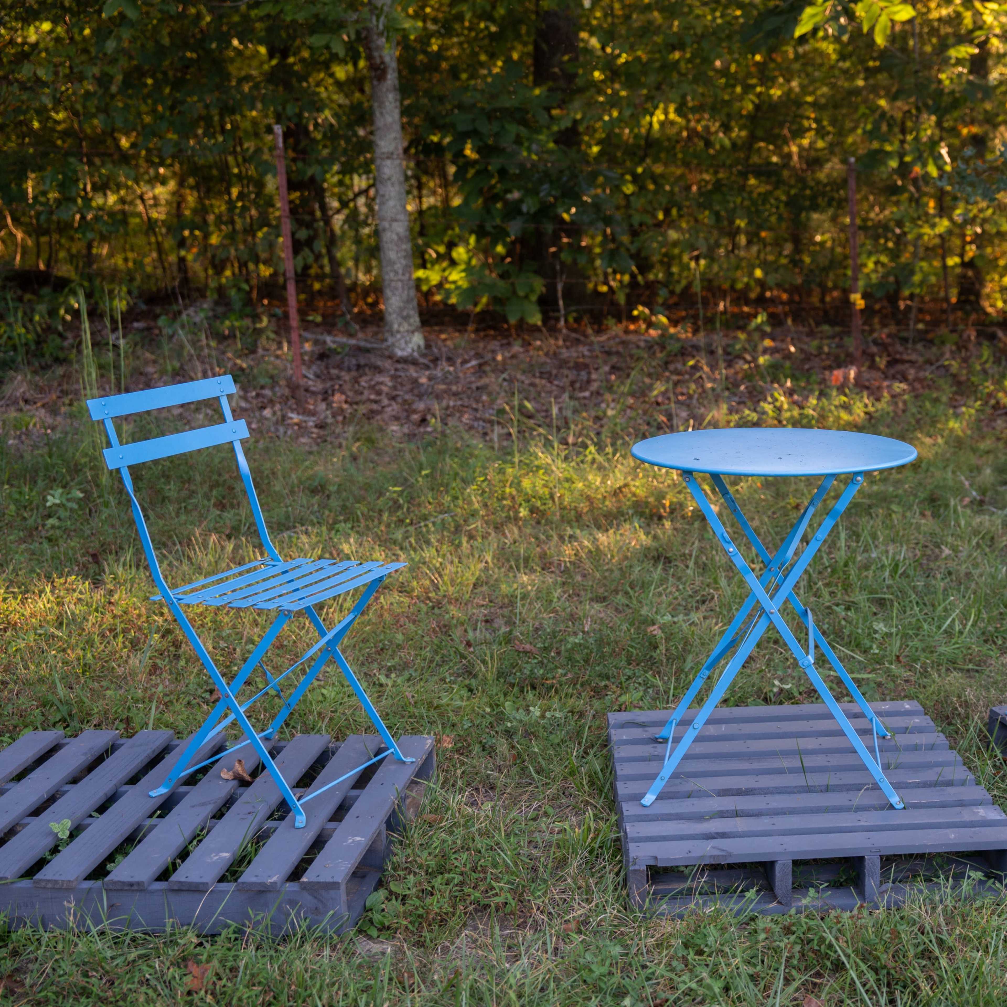 Two blue metal chairs are positioned around a small blue round table on wooden pallets in a grassy area surrounded by trees.