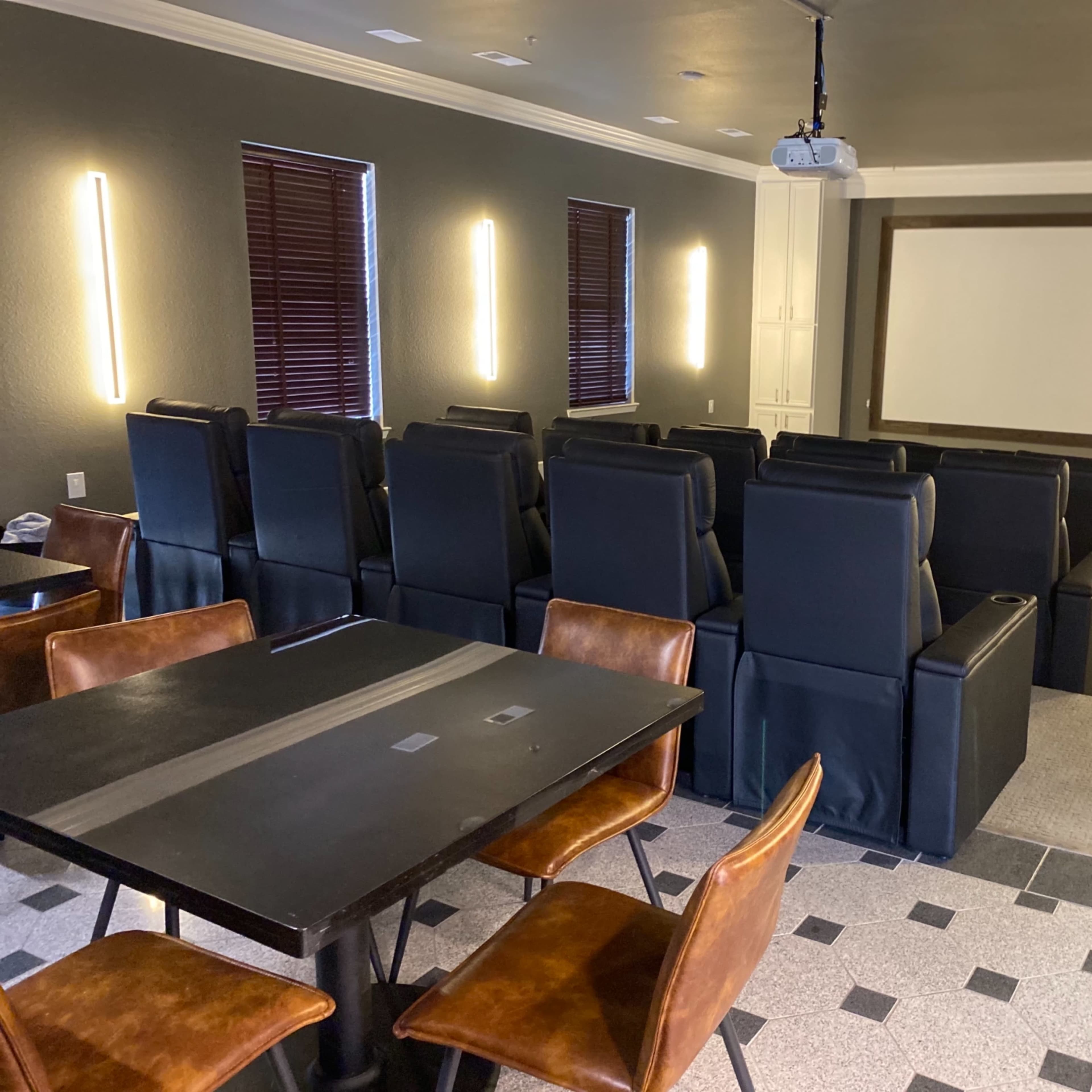A home theater with black reclining chairs arranged in rows, a large projection screen, and a table with wooden chairs in the foreground.