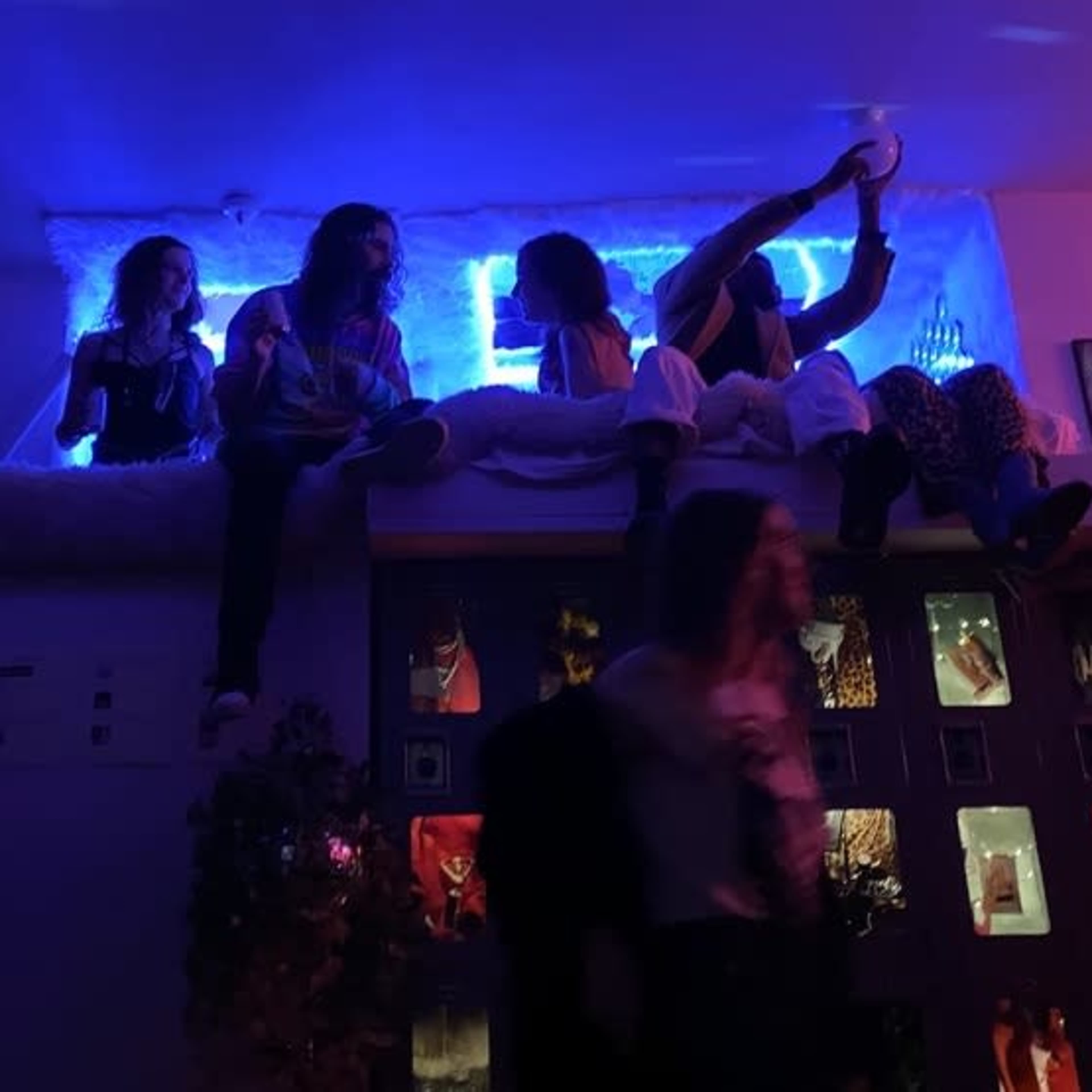 A group of people sits on a bed and floor in a dimly lit room with colorful lighting and decorative elements.