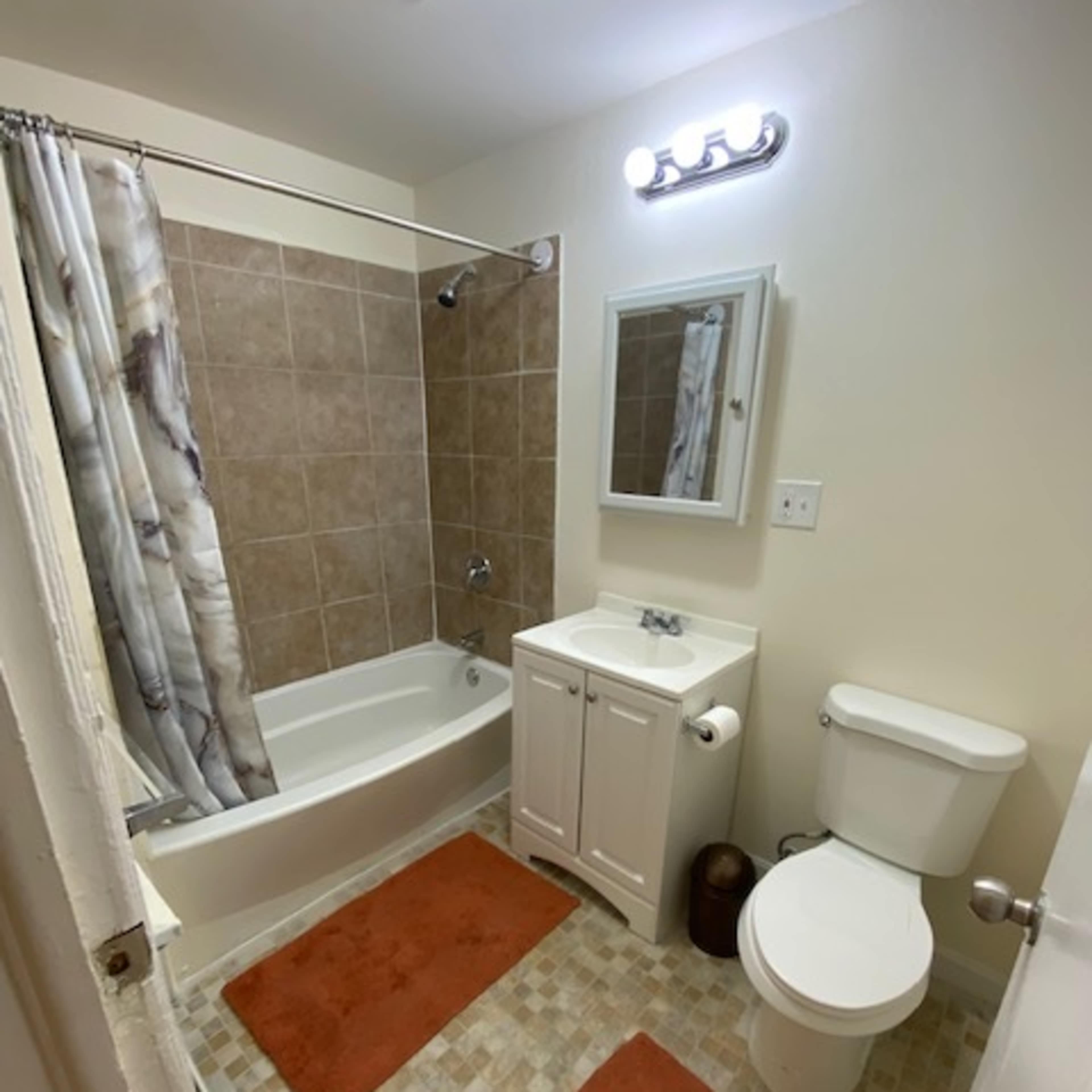 A small bathroom with a bathtub, a toilet, a sink with a mirror, and two orange bath mats on the tiled floor.