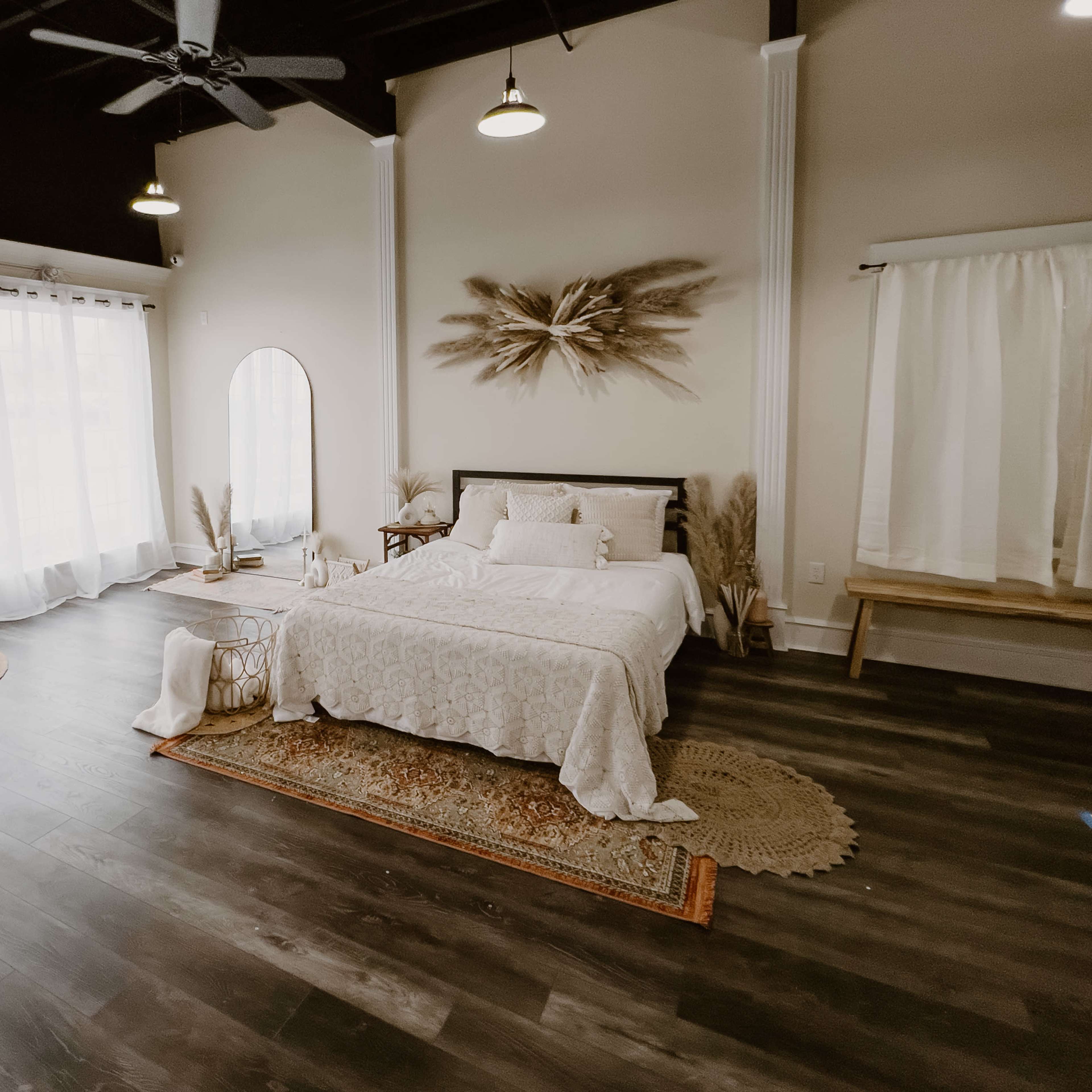 A well-lit bedroom with a bed, white bedding, large windows, and decorative elements like a mirror and woven baskets.