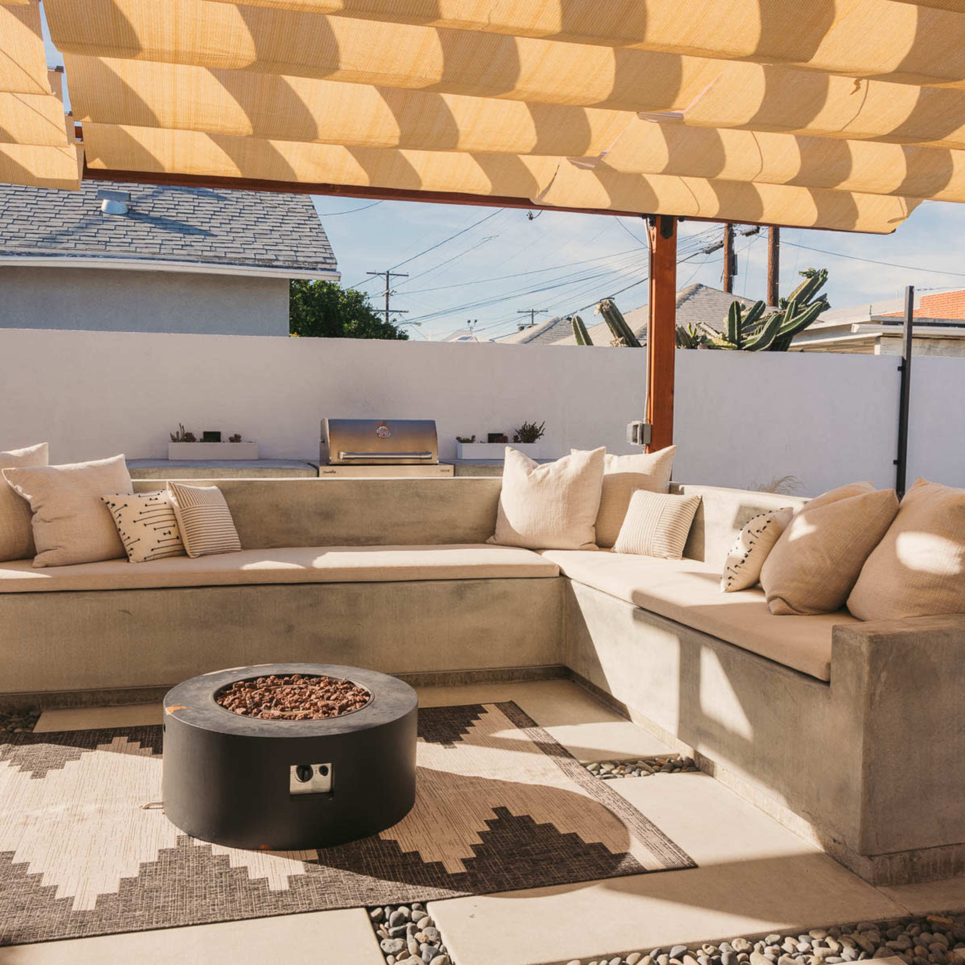 The scene features a modern outdoor seating area with a concrete sectional couch, a fire pit, and a large shaded pergola.