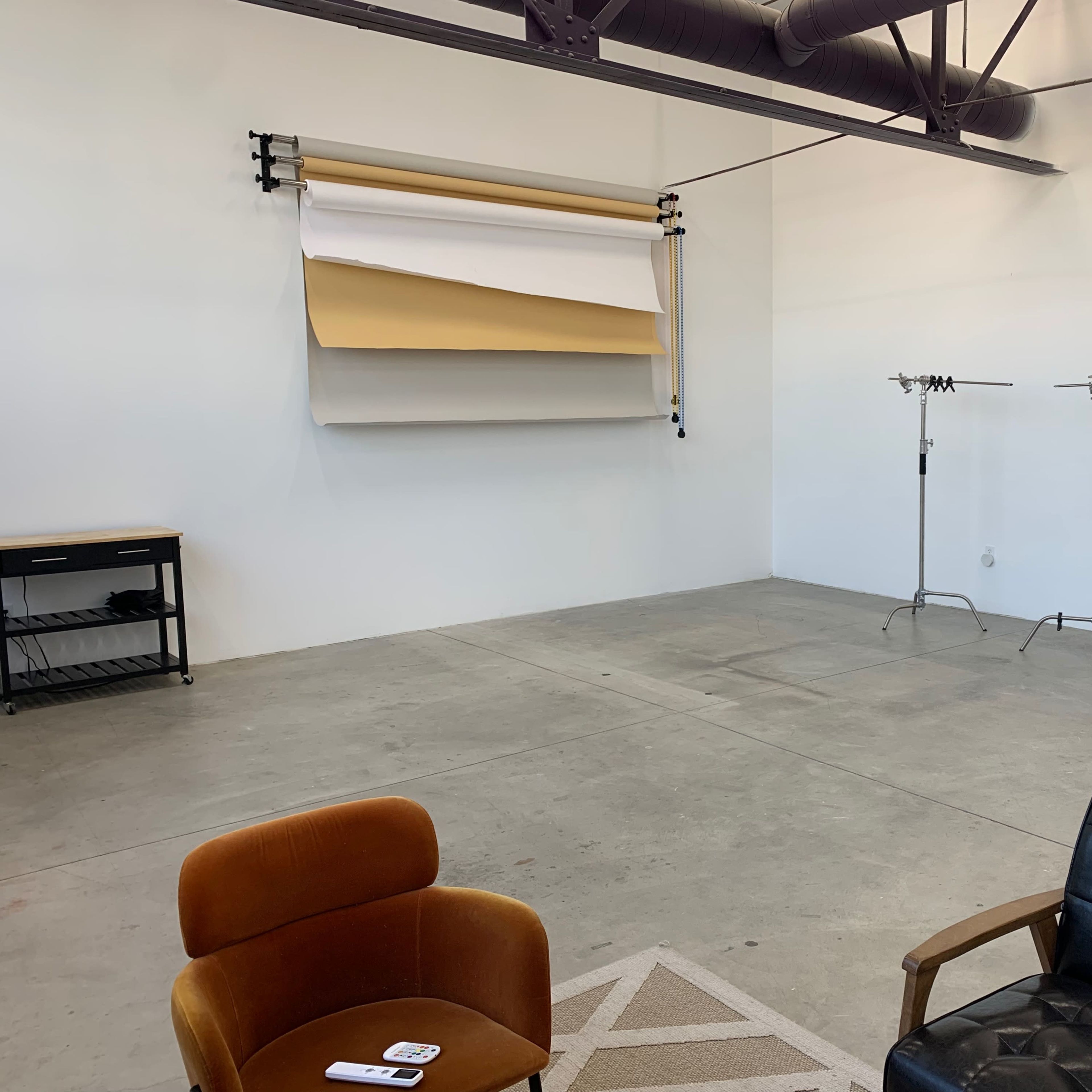 A spacious room with concrete flooring, featuring a wall-mounted backdrop, two standing clothing racks, and a couple of seating options.