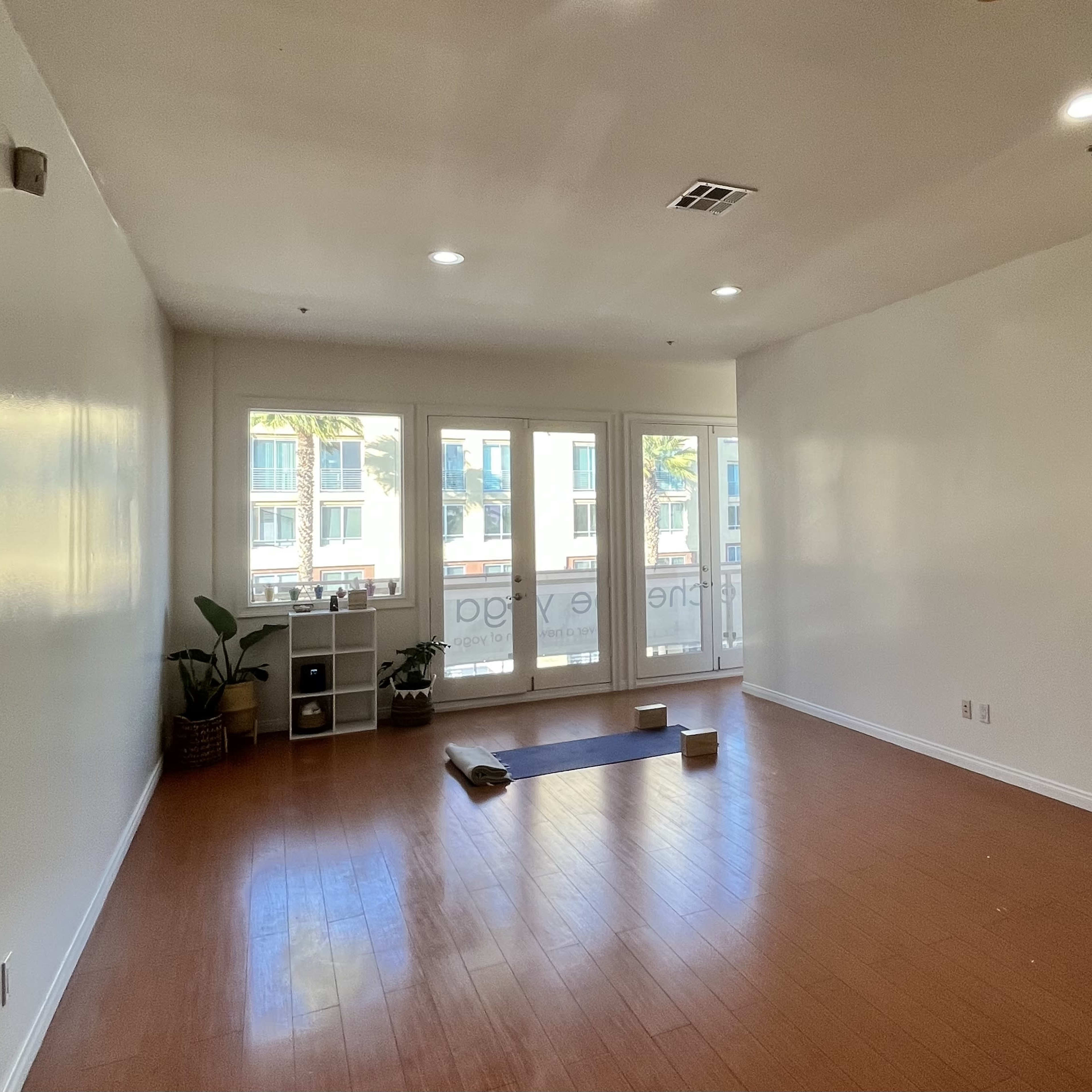A spacious, empty room with wooden flooring, a yoga mat, and two yoga blocks, featuring large windows that allow natural light and view of palm trees outside.