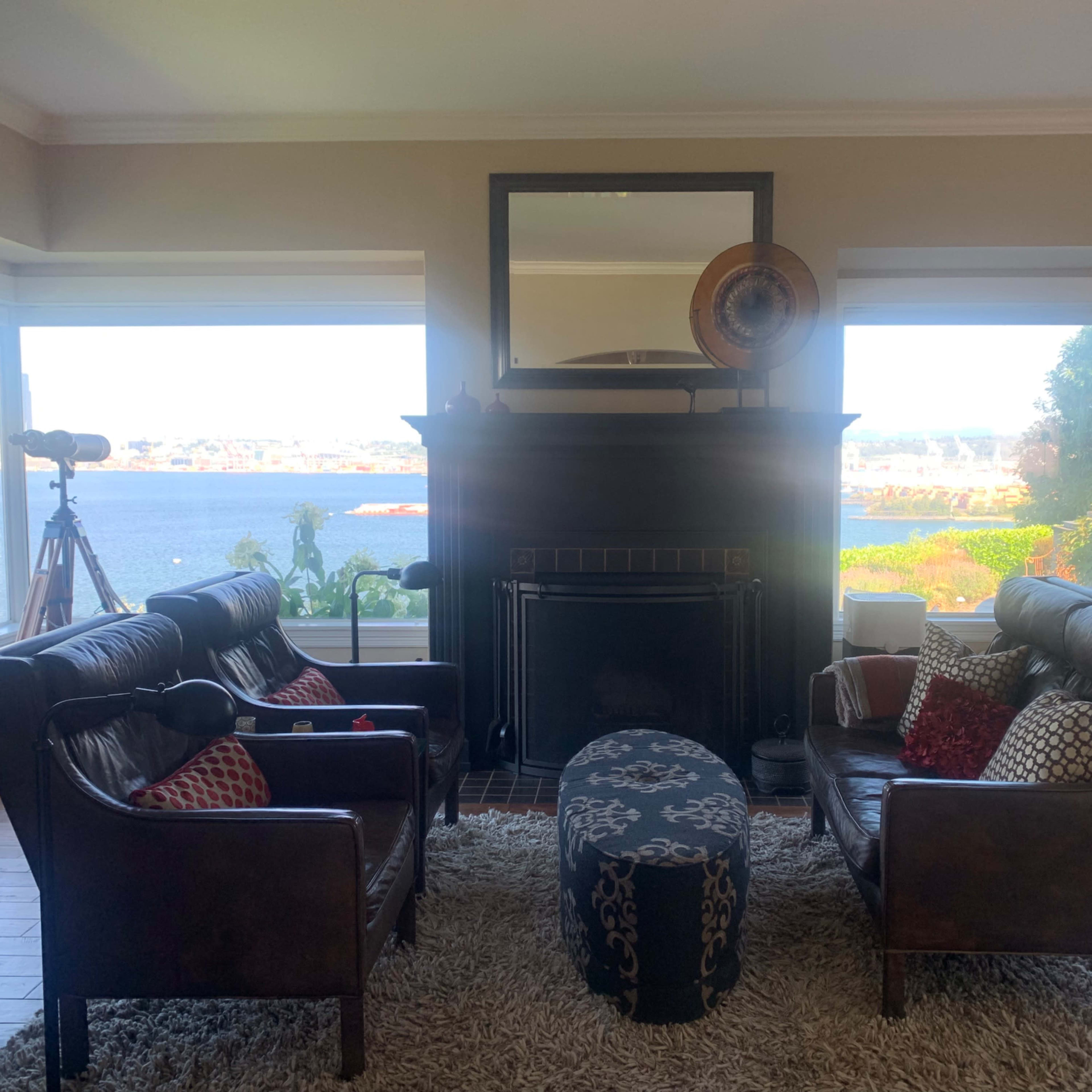 A living room with two leather armchairs facing a fireplace, a round ottoman in the center, and large windows overlooking a body of water.