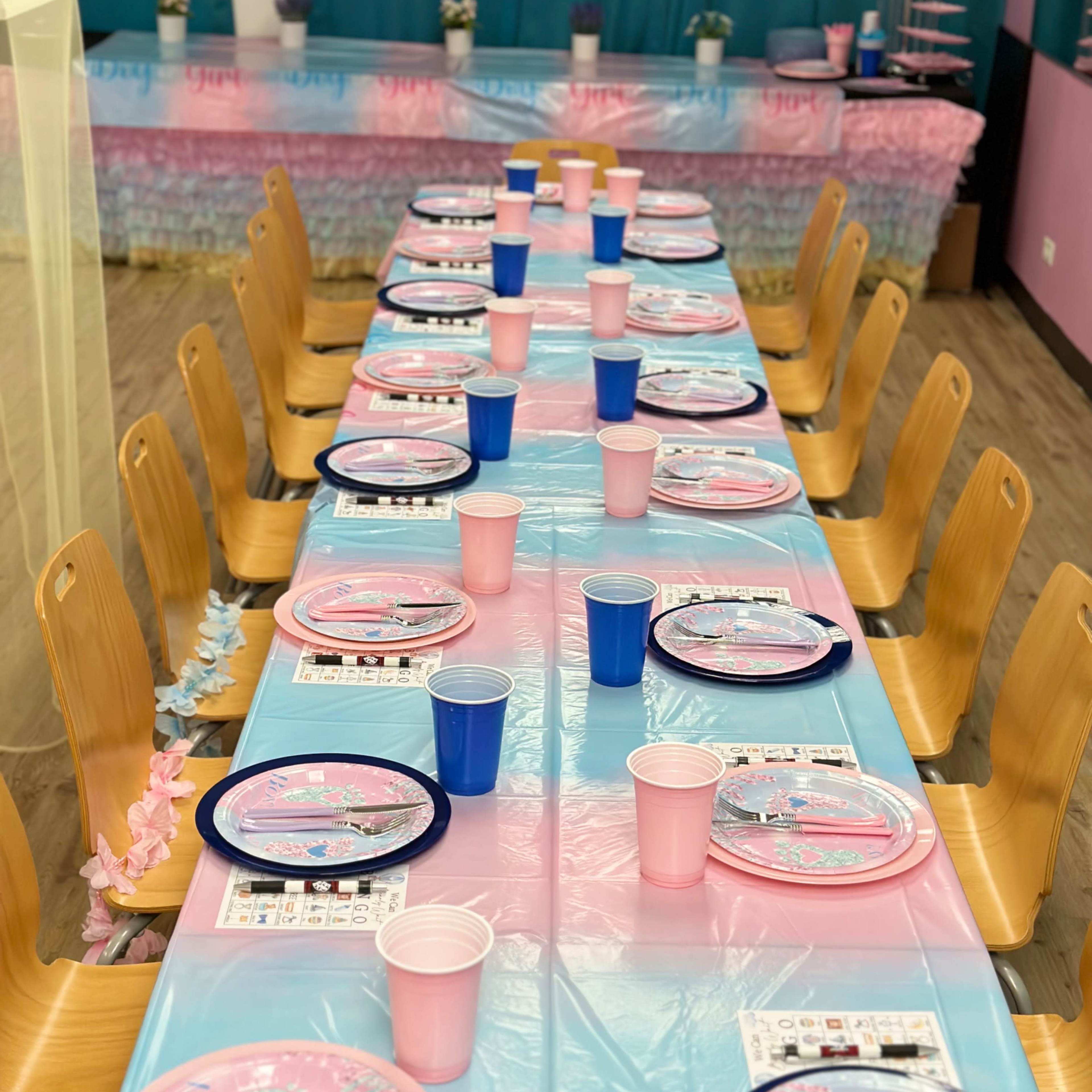 A long table is set with blue and pink plates and cups, topped with a teal and pink tablecloth, in a decorated room.