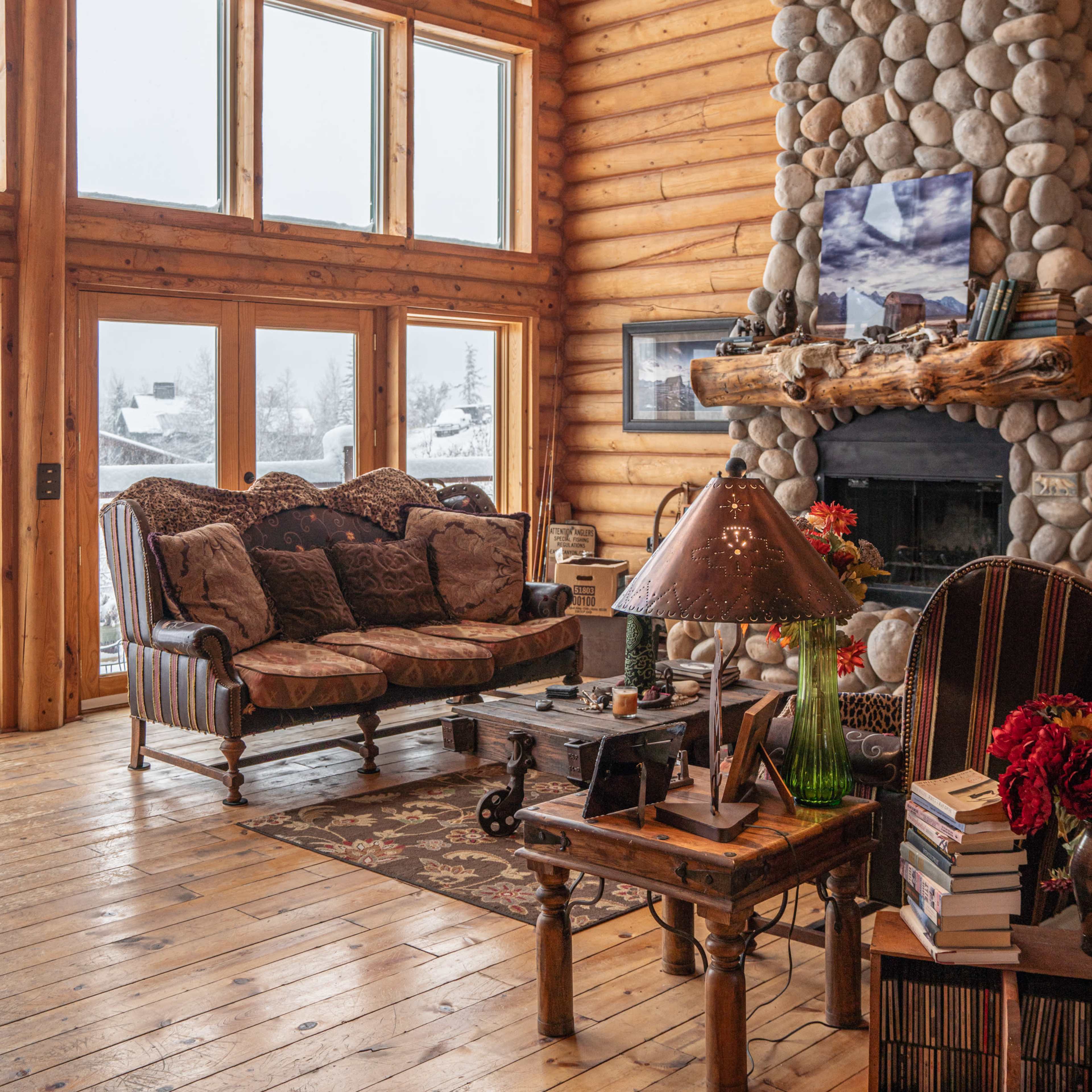 A cozy wooden living room features large windows, a stone fireplace with a mounted deer head, and rustic furniture.