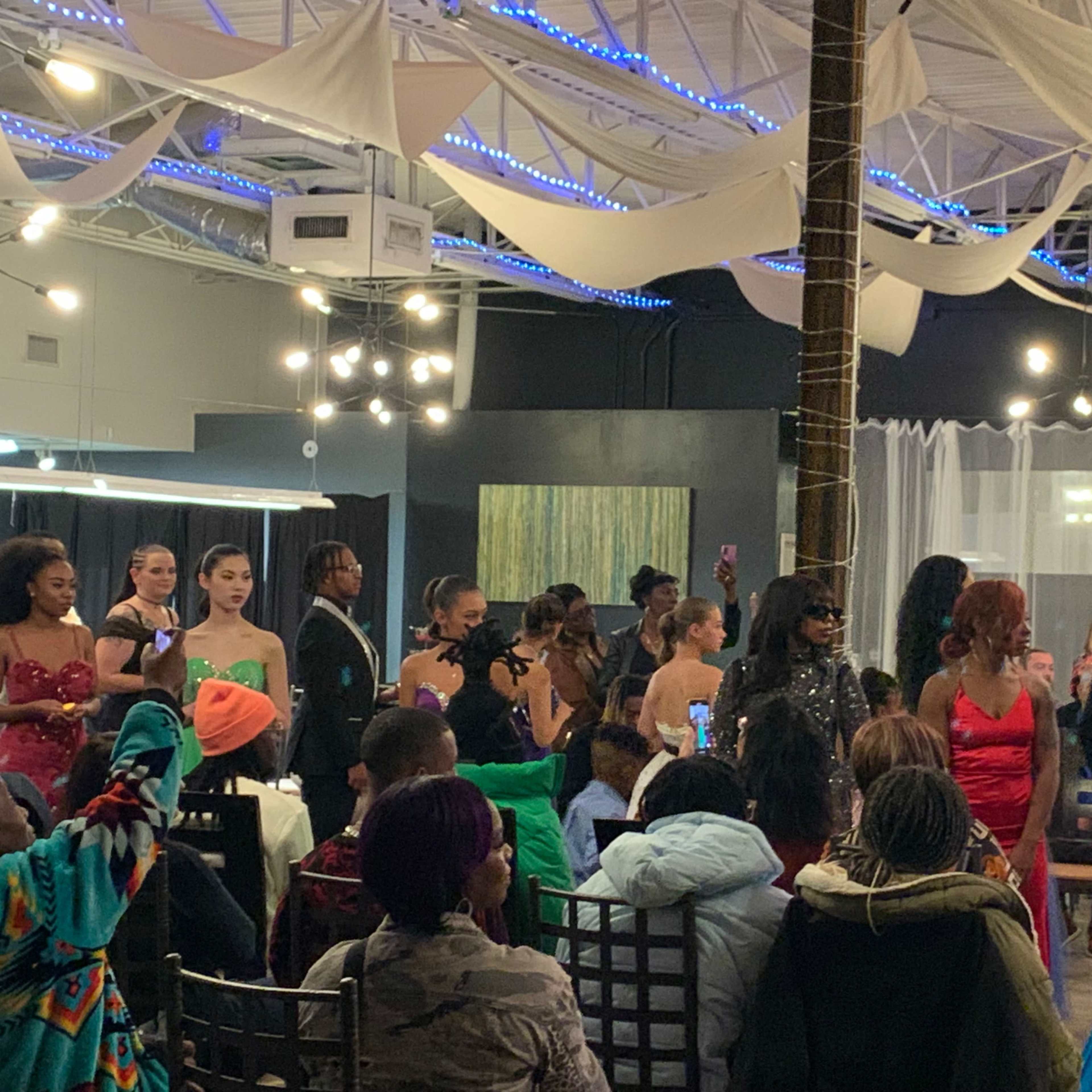 A group of people dressed in formal attire gathers in a decorated indoor space for an event, with some participants displayed on a small stage.