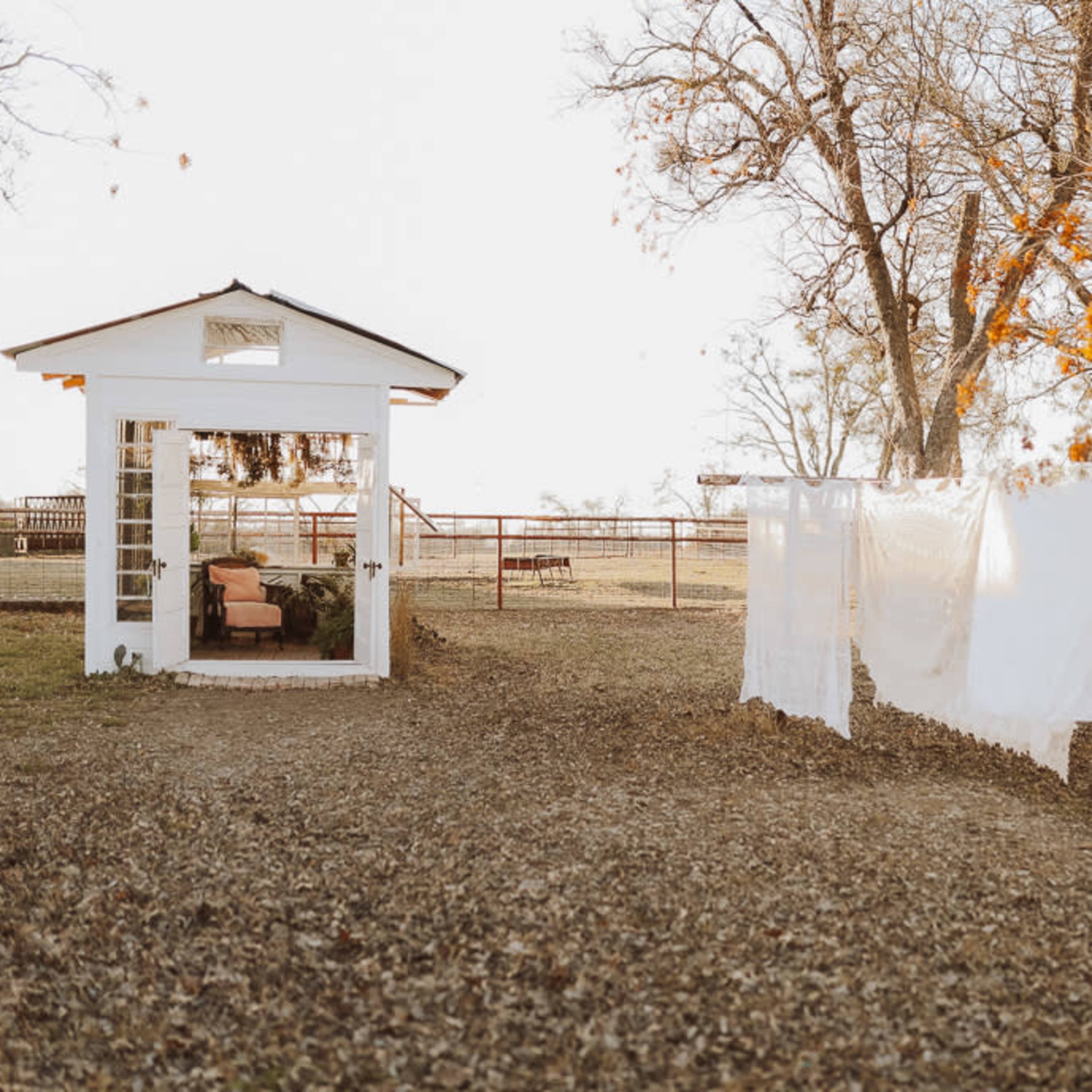 A small white structure with large windows stands beside a clothesline holding freshly laundered white sheets in a grassy area surrounded by trees.