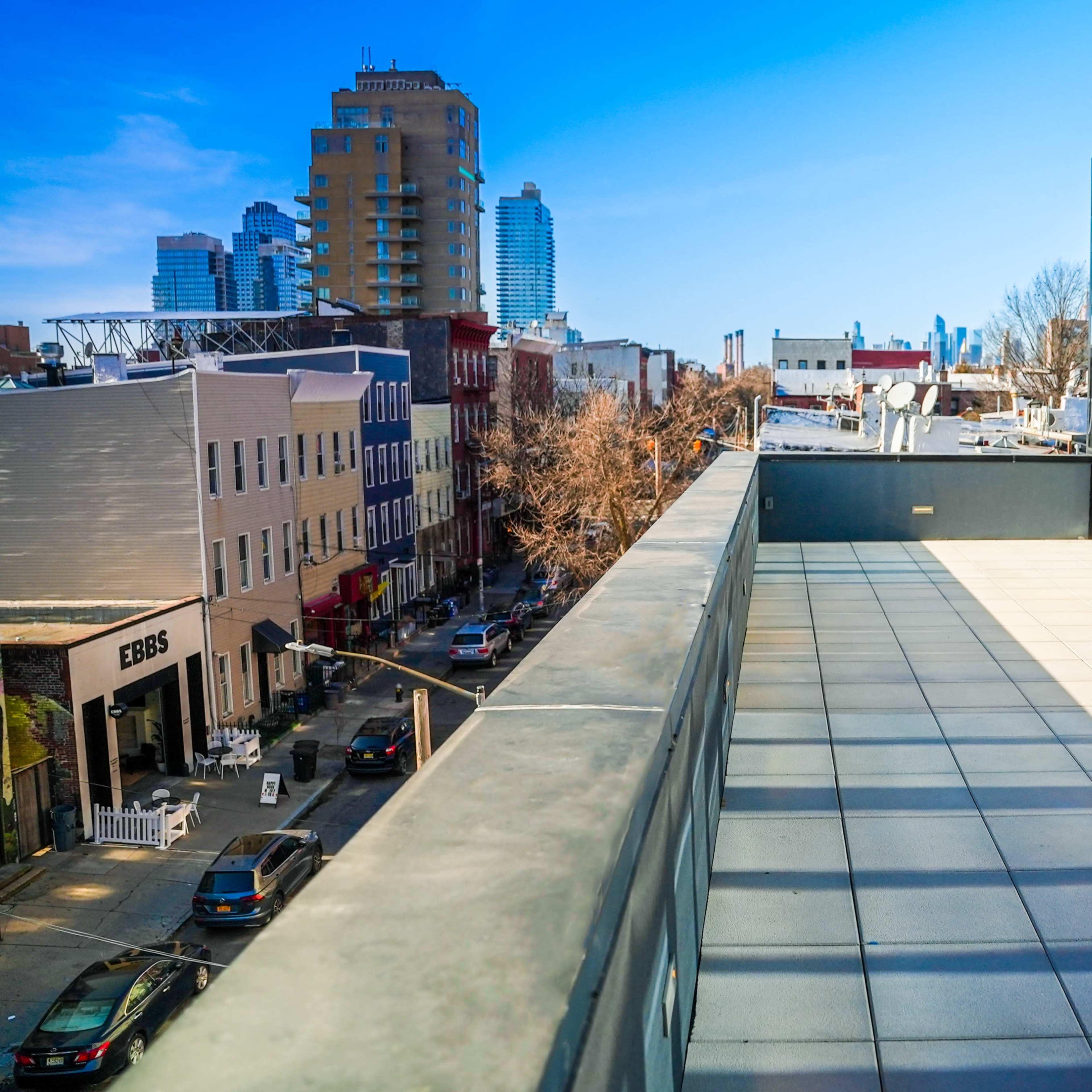 A rooftop view overlooking a street with several buildings and parked cars under a blue sky.