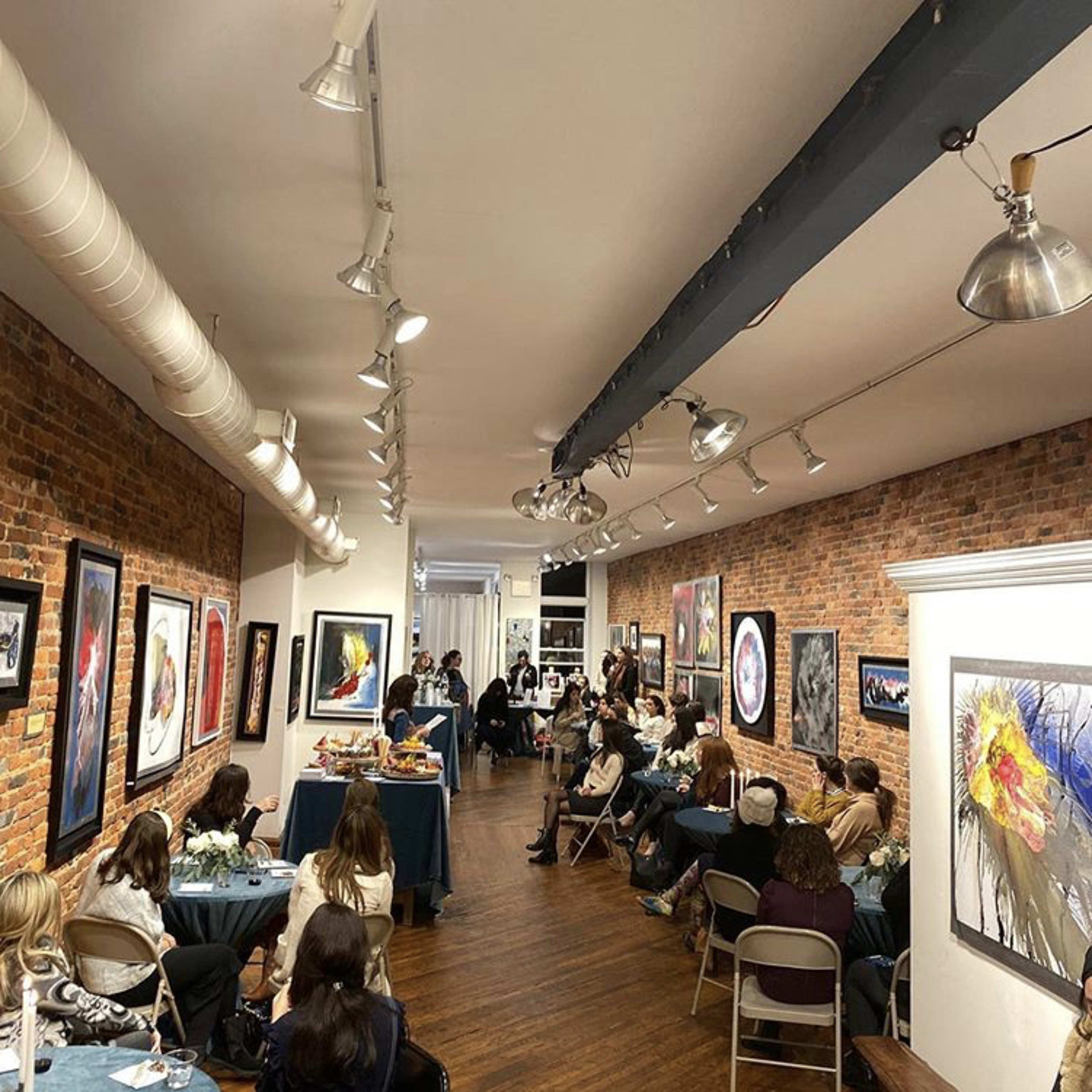 A group of people sits in a gallery with brick walls, surrounded by various framed artworks, while an event takes place at the front of the room.