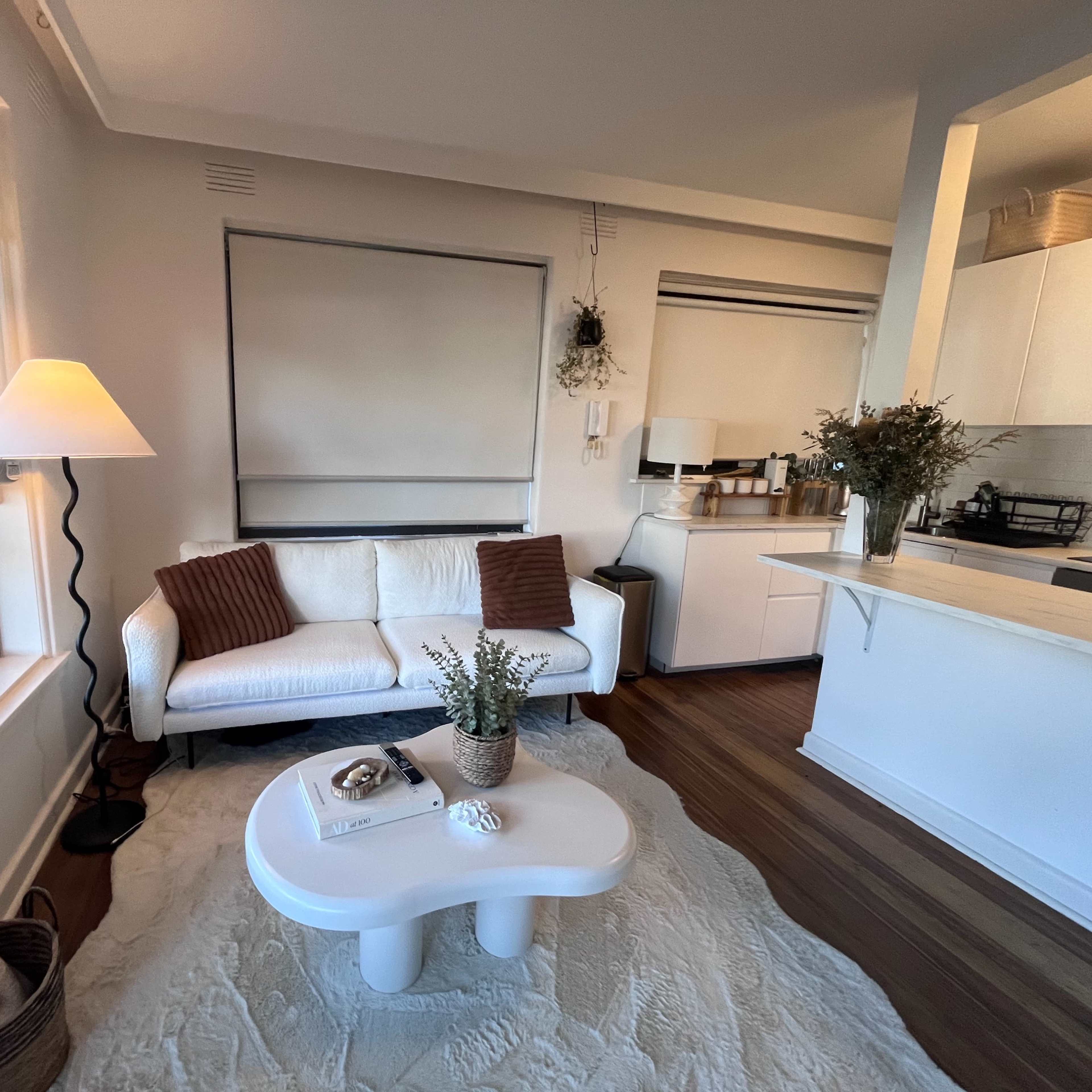 A living room with a white couch, a round coffee table, a floor lamp, and a kitchen area in the background.