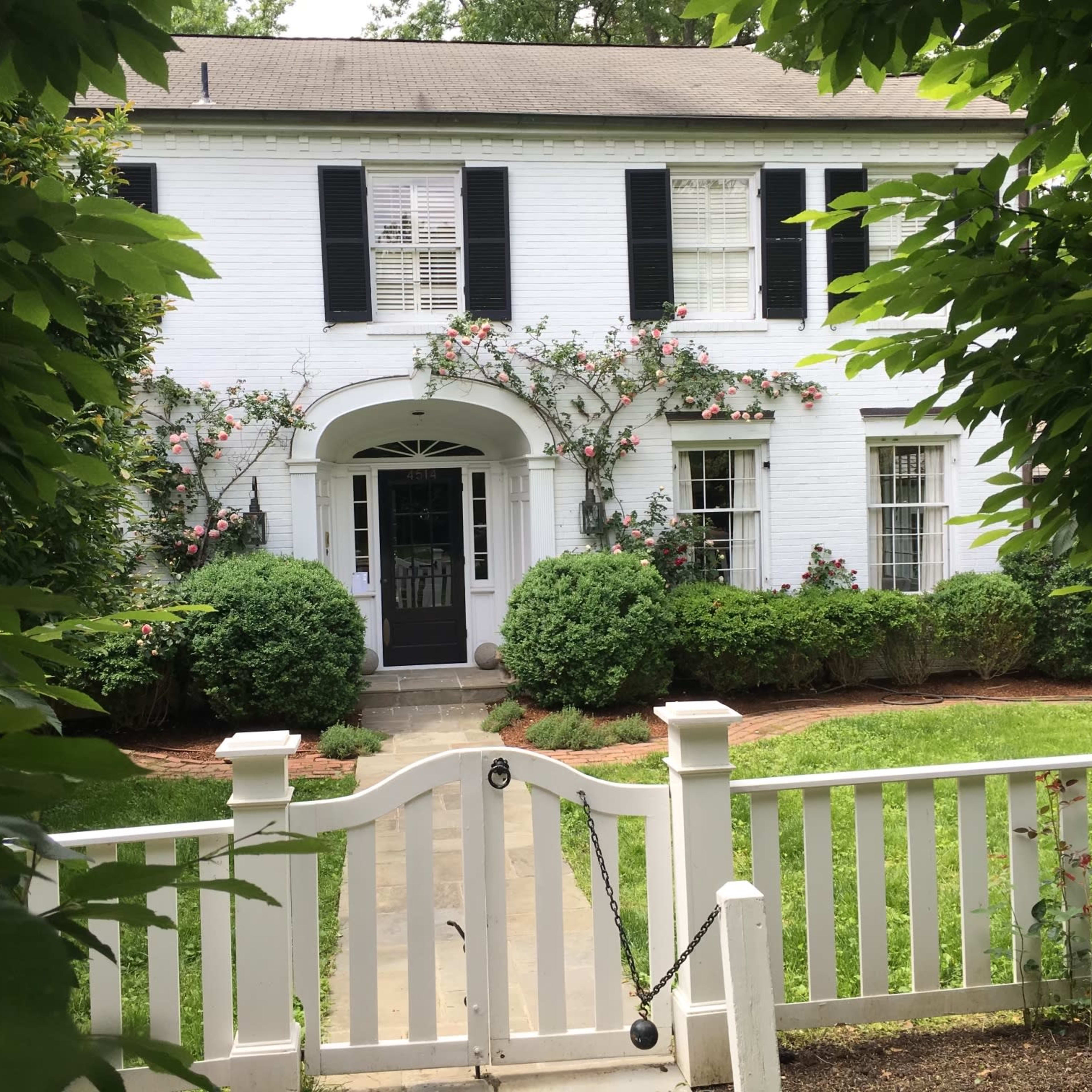 A white brick house with black shutters is framed by lush greenery and roses, featuring a covered front entrance and a white picket fence.