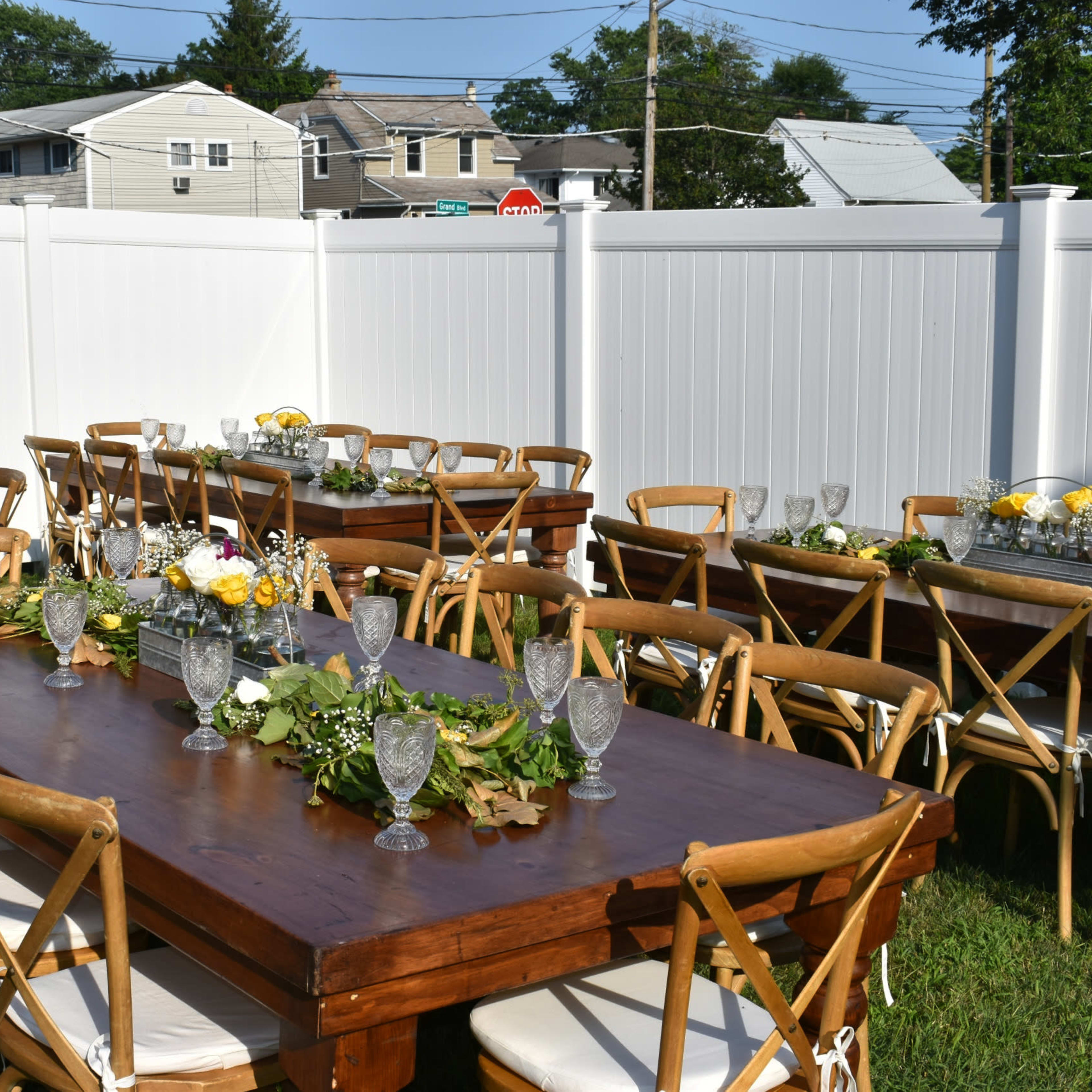The scene features two long wooden tables set outdoors, adorned with glassware and floral centerpieces, surrounded by wooden chairs in a grassy area enclosed by a white fence.
